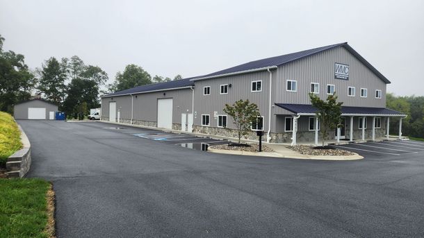 A long, grey metal commercial building with a black roof, stone foundation, and asphalt parking lot on a cloudy day.