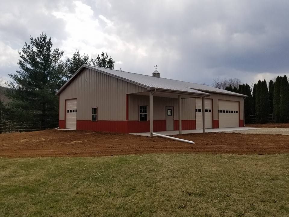 A beige and red metal pole barn building with three garage doors and a covered side porch sits on a graded dirt lot.