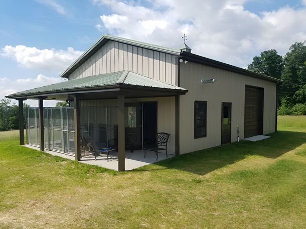 A tan metal building with a green roof, featuring a covered porch with wire enclosures, set in a grassy field.