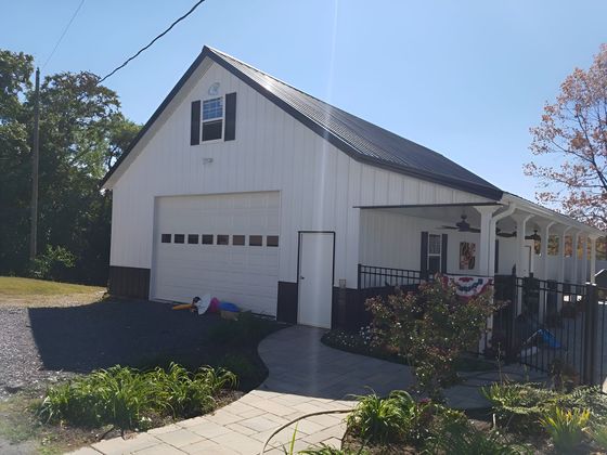 A white, gable-roof garage with a side porch and a dark roof, set on a gravel lot with a paved walkway in the foreground.
