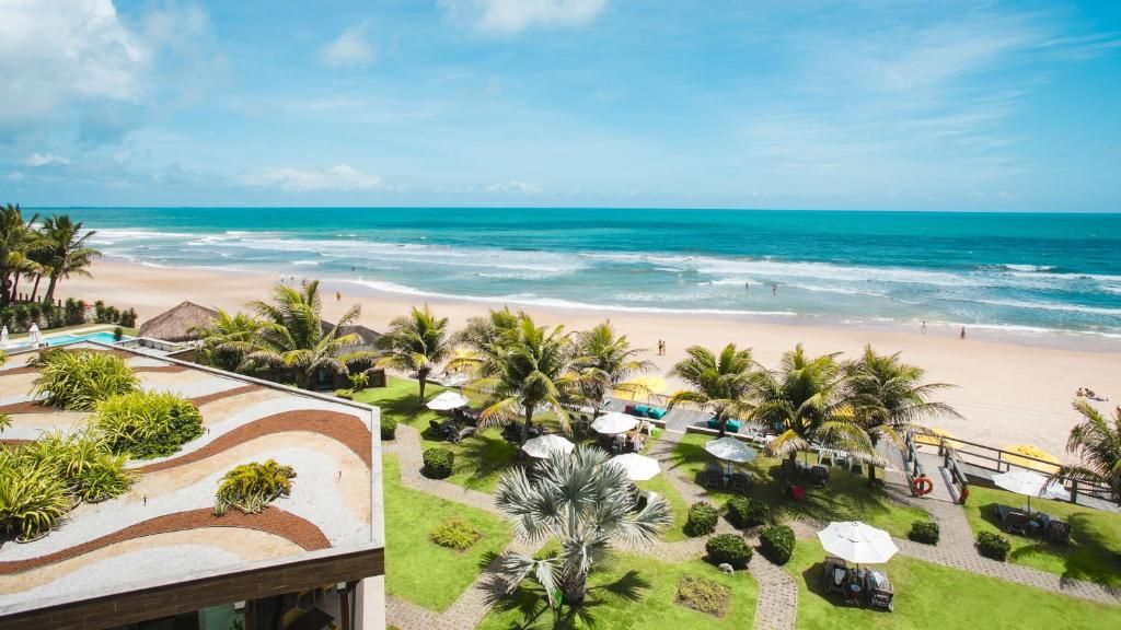 Um artista barbudo com óculos escuros e camiseta branca posa em frente a um prédio. Na fachada, há um grande mural em preto e branco com detalhes em laranja, retratando uma figura feminina estilizada.