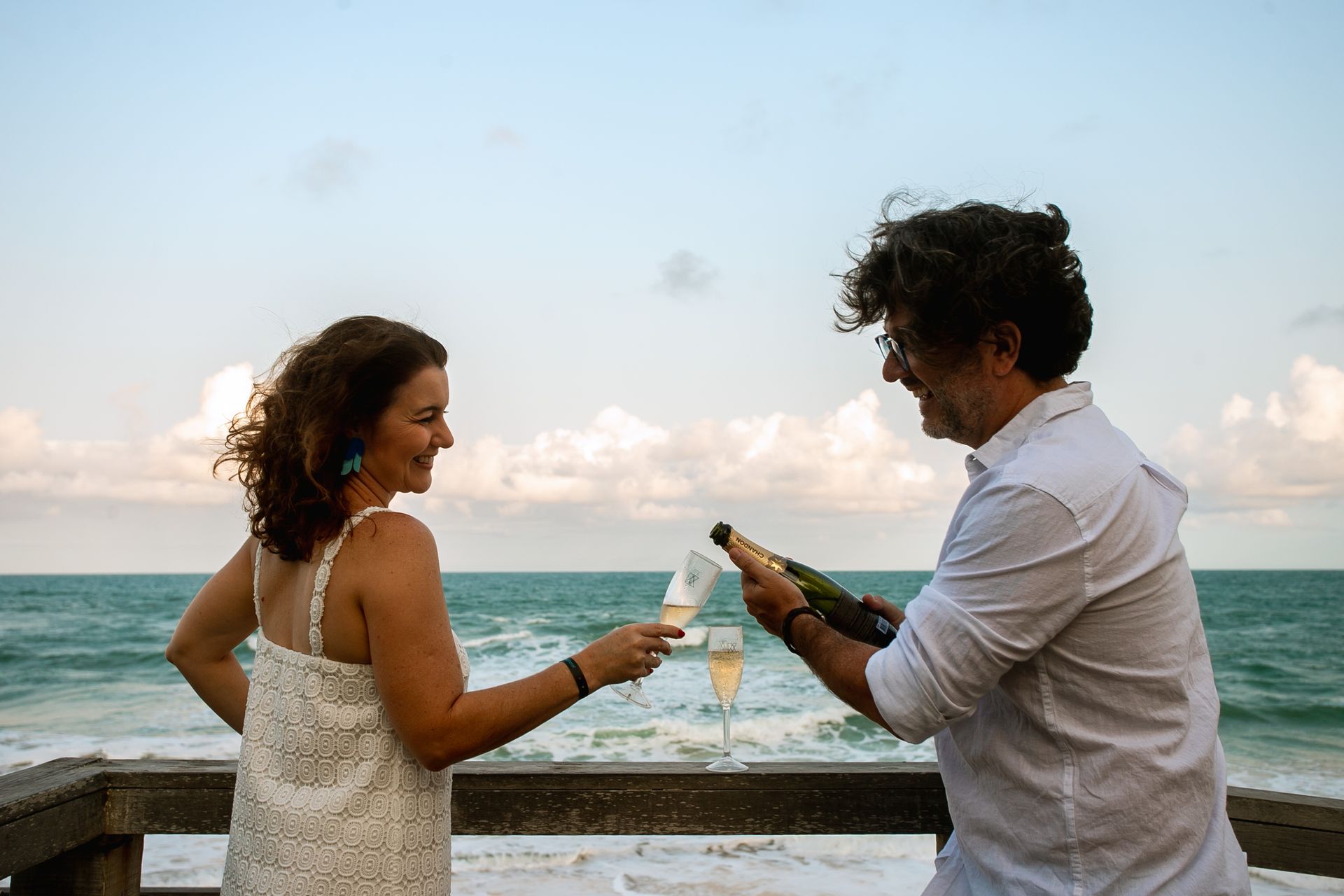 Um casal sorridente em um deque de madeira diante do mar; o homem serve espumante em uma taça enquanto a mulher segura a dela para brindar ao entardecer.