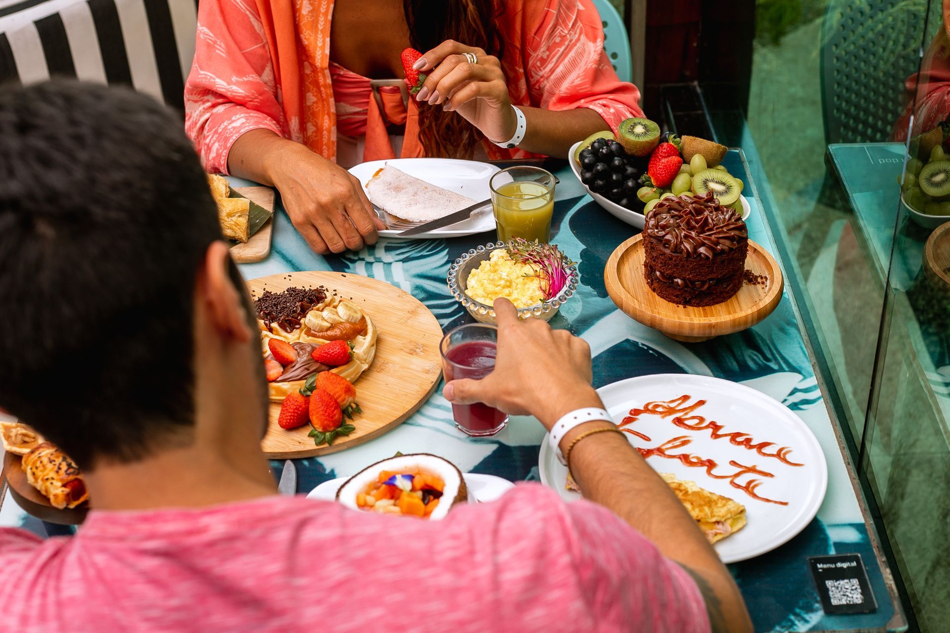 Um casal desfrutando de um café da manhã em uma mesa, repleta de diversas opções. Com frutas frescas, sucos, um bolo de chocolate, waffle com morangos, uma tapioca e uma omelete.