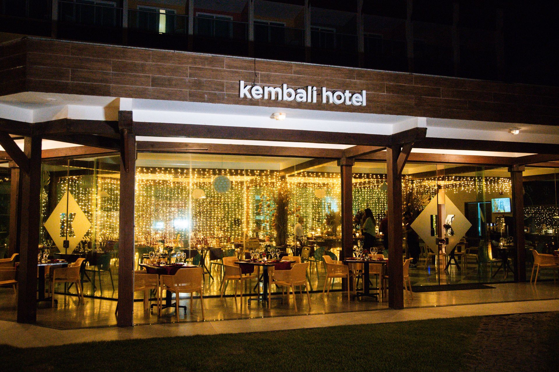 Fachada iluminada do restaurante do Kembali Hotel à noite, com decoração de luzes douradas, mesas postas para jantar e estrutura de madeira ao redor do espaço envidraçado.