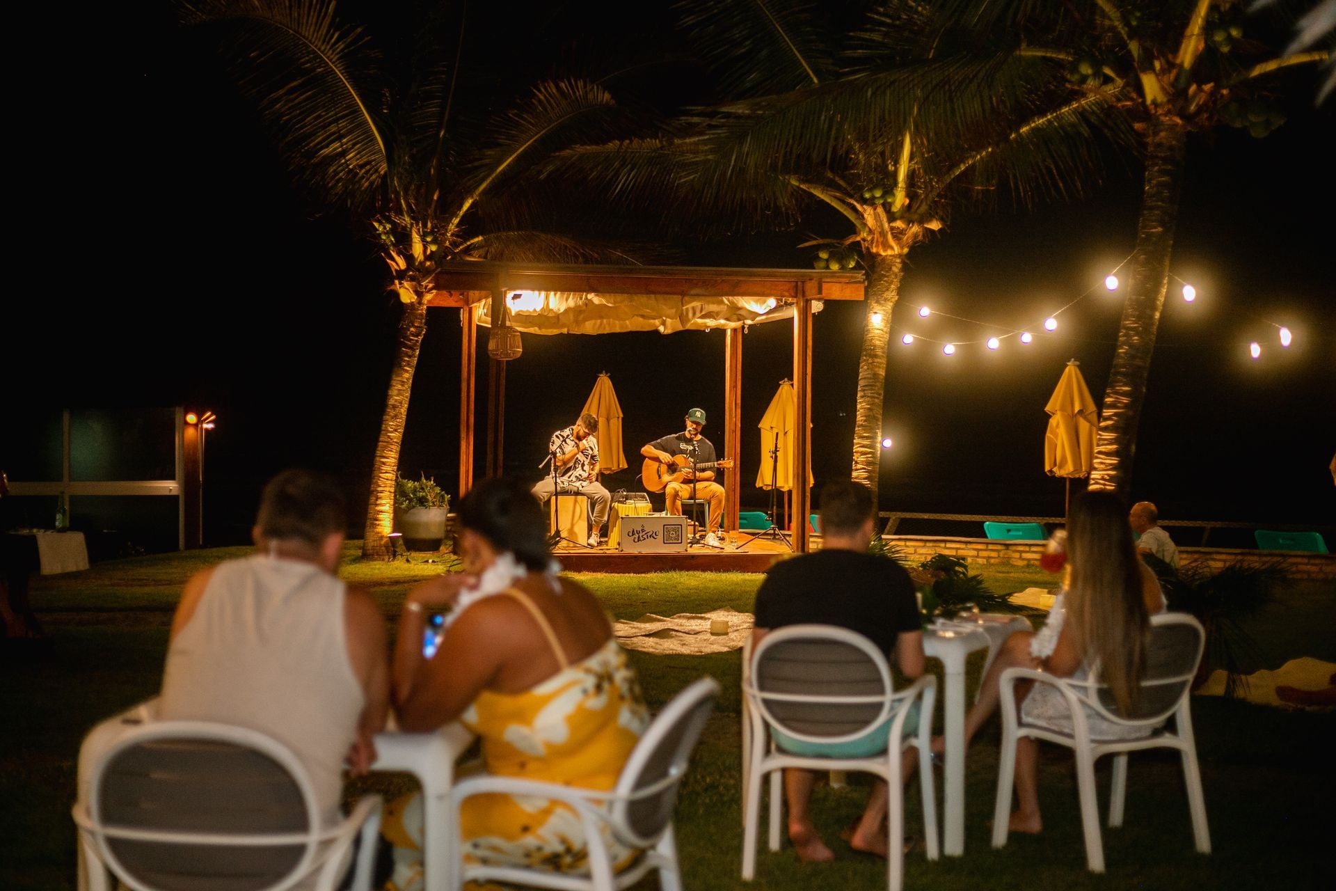 pessoas sentadas assistindo música ao vivo em área externa à noite com palco, coqueiros e iluminação decorativa