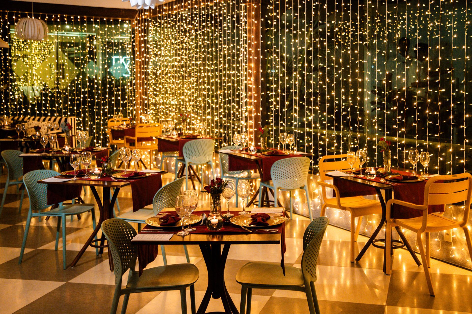 Sala de jantar decorada com luzes douradas, mesas arrumadas para um jantar especial e cadeiras coloridas em um ambiente acolhedor e festivo.