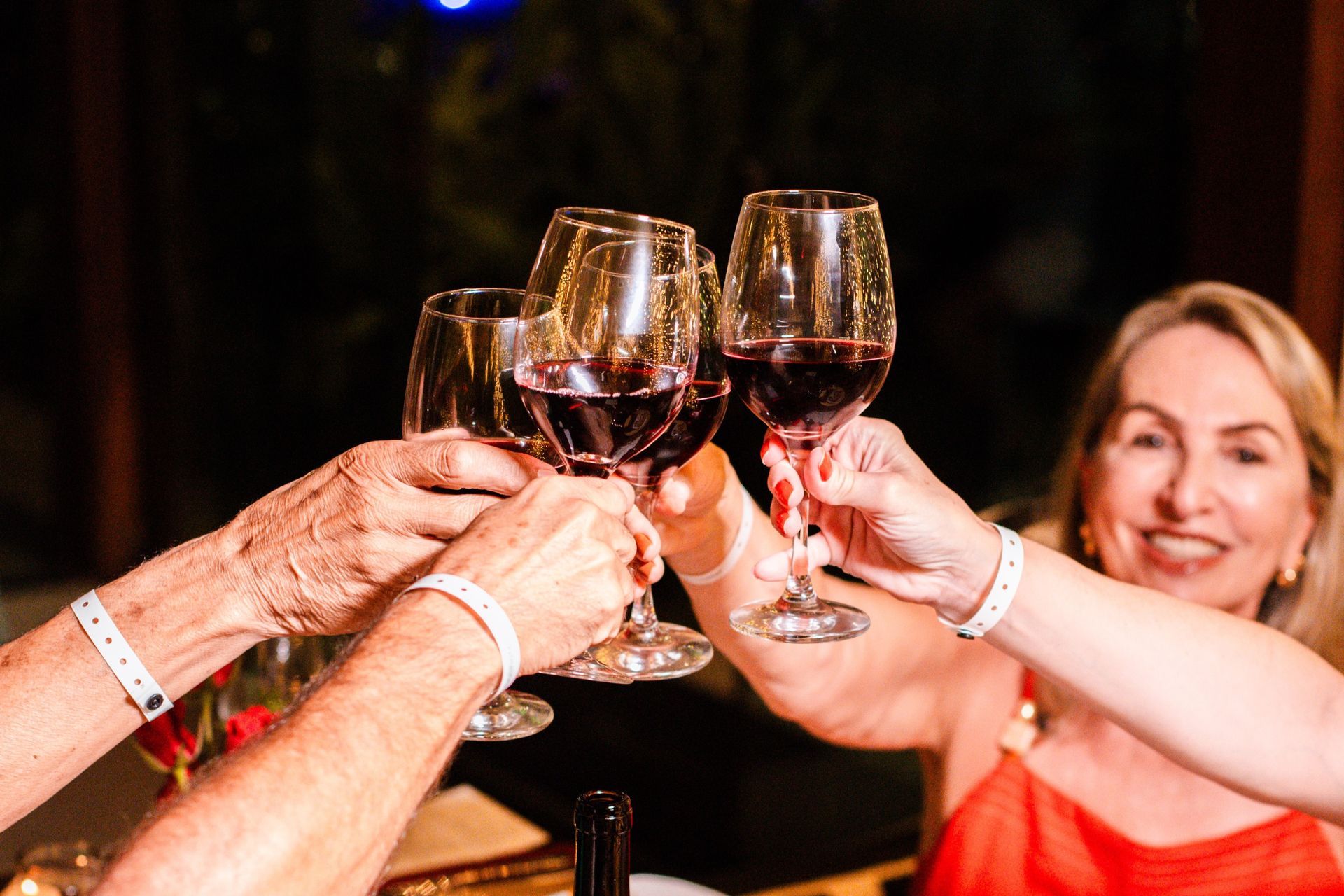 Grupo de pessoas brindando com taças de vinho tinto em um ambiente festivo, com mãos erguidas e clima de celebração.