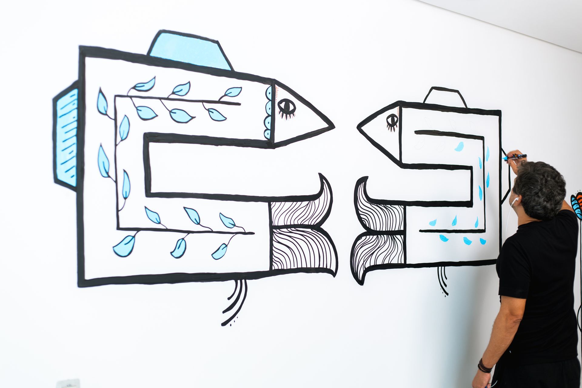 Um homem, visto de costas, pintando um mural minimalista de dois peixes estilizados em uma parede branca. Os peixes são formados por linhas grossas pretas em forma de labirinto, com barbatanas azuis e detalhes internos. O peixe maior tem folhas azuis desenhadas e o artista está trabalhando no peixe menor, adicionando gotas azuis.