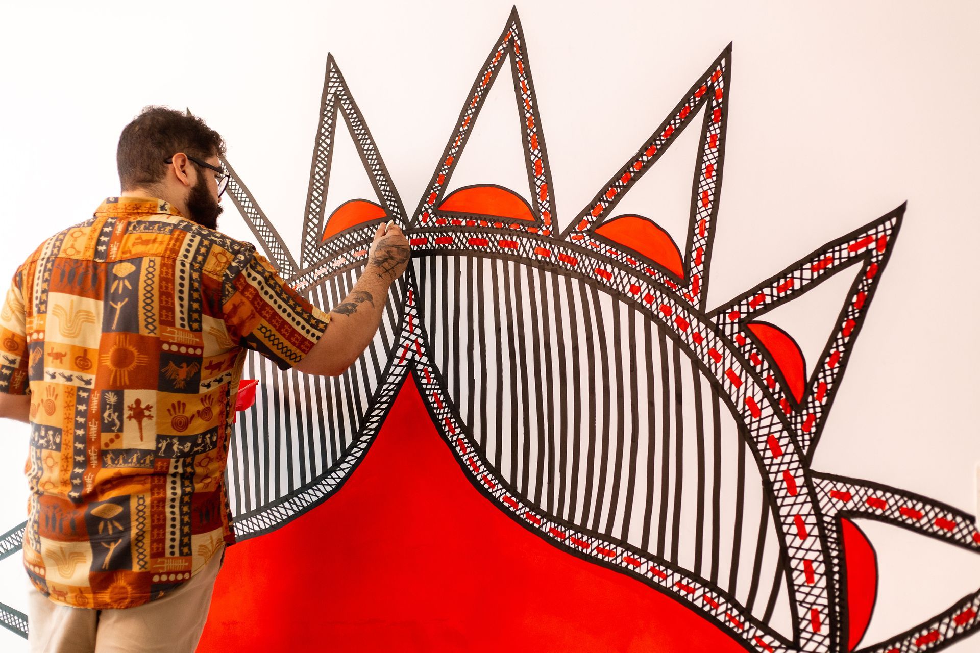 Artista (homem barbudo) em uma camisa estampada, pintando um grande mural. Ele está de costas, aplicando linhas pretas e padrões pontilhados em formas que lembram símbolos tribais em vermelho vibrante. A imagem capta o artista no processo focado de criação de uma obra de arte urbana detalhada.