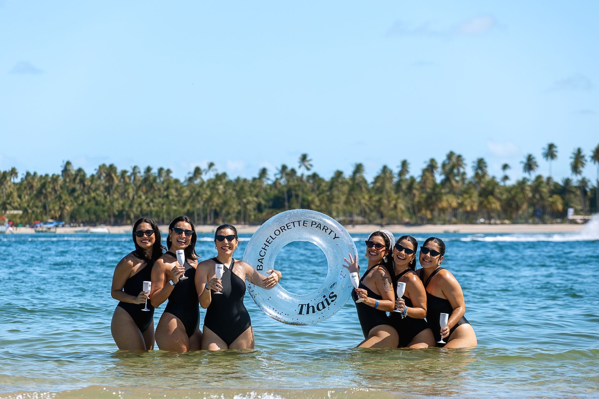 Seis mulheres de maiô preto, no mar de água rasa, segurando uma boia transparente.