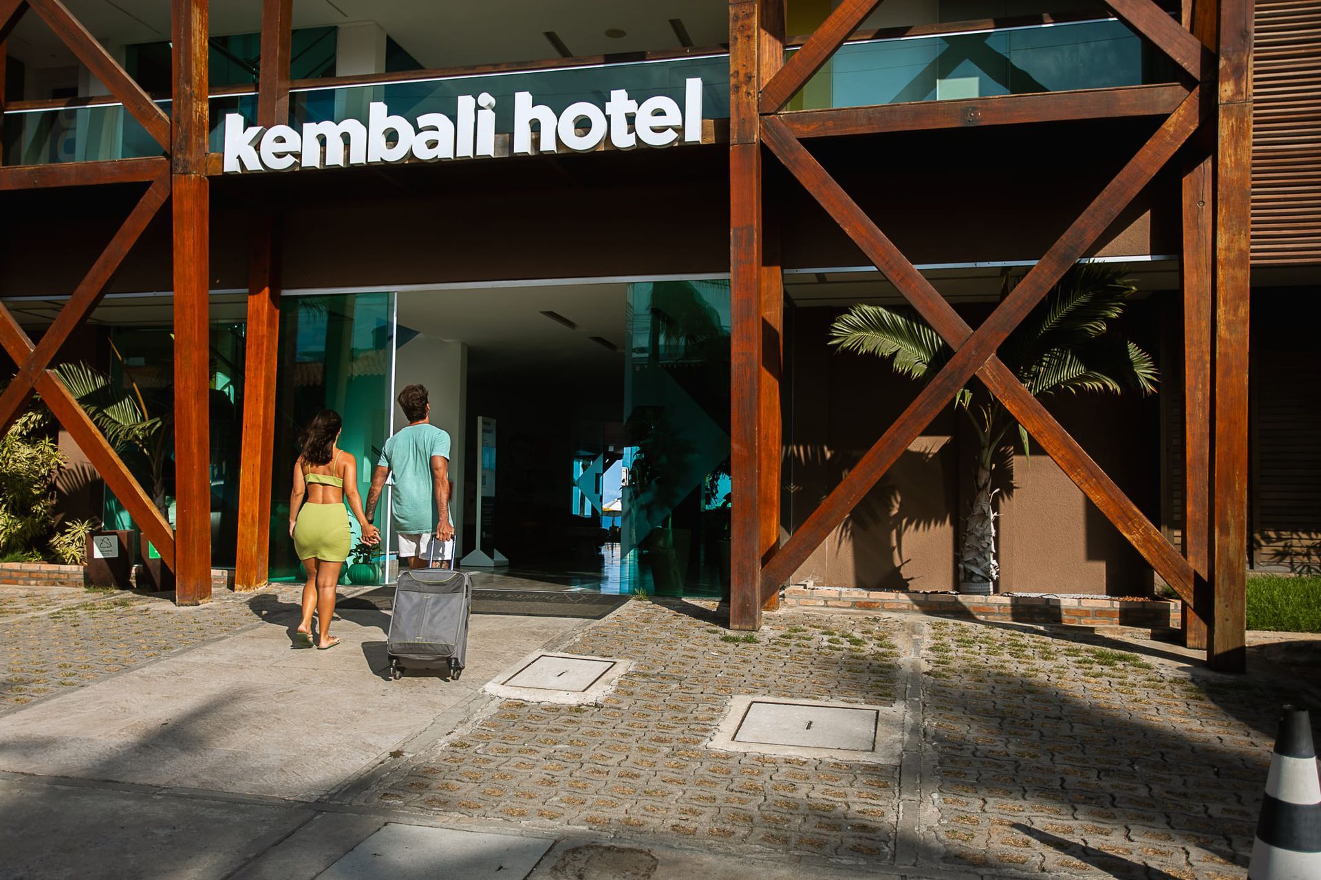 Entrada do hotel Kembali, com fachada moderna em madeira e vidro. Um casal caminha em direção à recepção; uma das pessoas puxa uma mala de rodinhas. O nome do hotel aparece em letras brancas acima da entrada, e há palmeiras e calçamento de pedra ao redor.