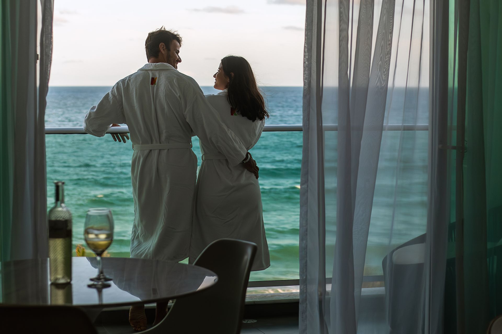 Casal abraçado na varanda do quarto do Kembali, com o mar ao fundo. Eles usam um roupão branco e estão sorrindo. Dentro do apartamento há uma mesa com uma garrafa de vinho branco e duas taças.