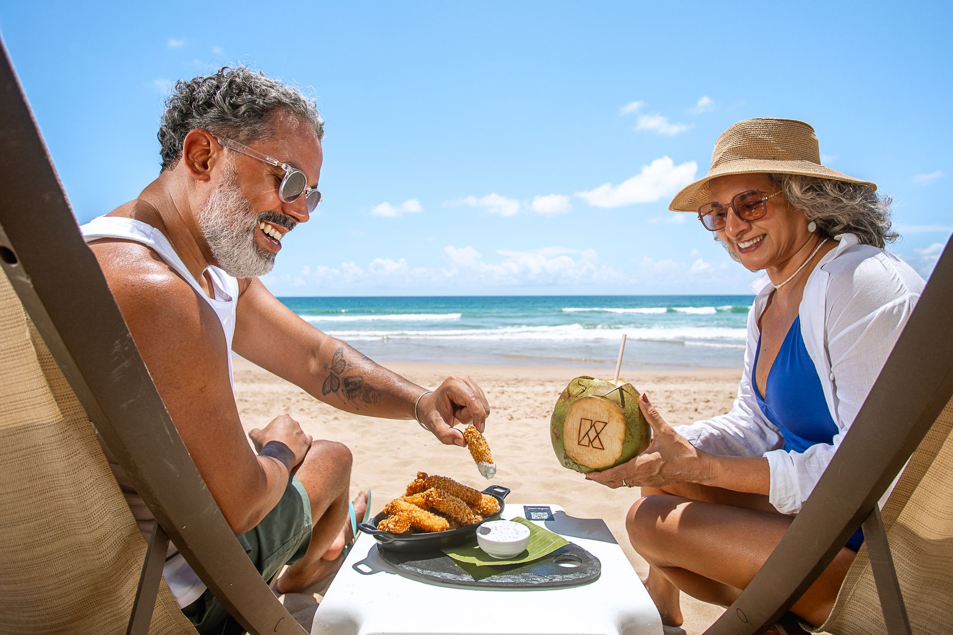Casal sorridente aproveitando o dia na praia, sentado em espreguiçadeiras à beira-mar. Eles compartilham um prato de petiscos e um coco gelado personalizado com a marca do hotel, em um cenário ensolarado com céu azul e mar ao fundo.