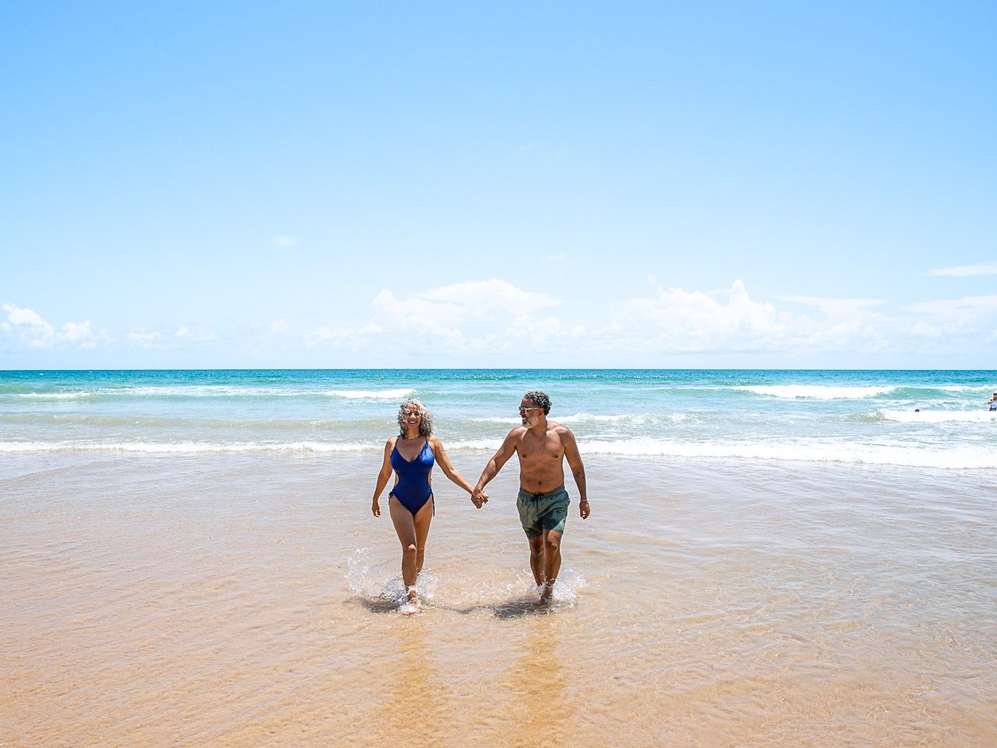 Um casal sorridente de meia-idade caminha de mãos dadas pela areia molhada de uma praia ensolarada, com o mar azul ao fundo.