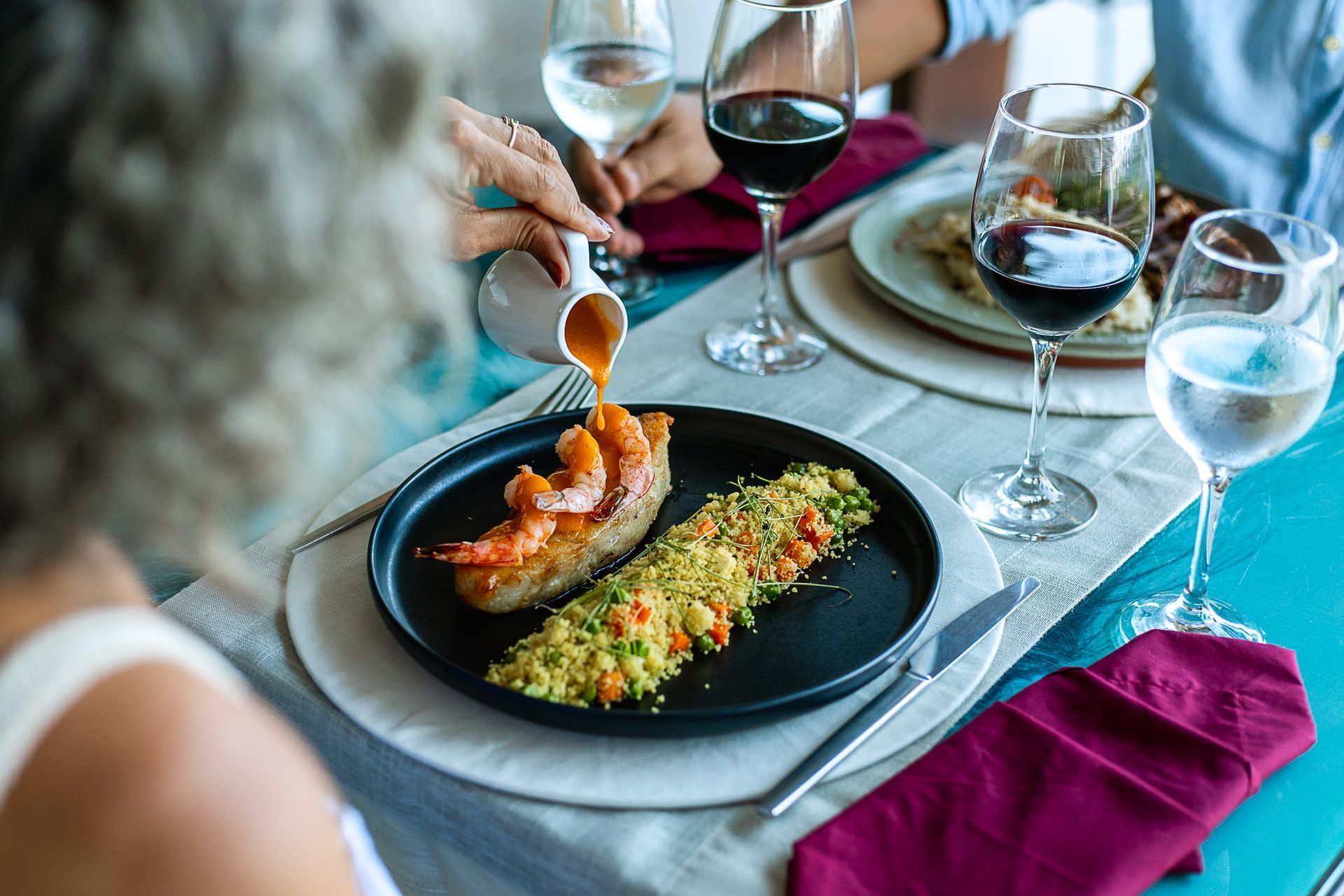 Mesa posta com taças de vinho e água, um casal sentado de frente para o outro, mas o foco são os pratos na mesa. O prato do primeiro plano é uma posta de peixe com camarões em cima e uma farofa ao lado. a Mulher derrama um molho laranja nos camarões.