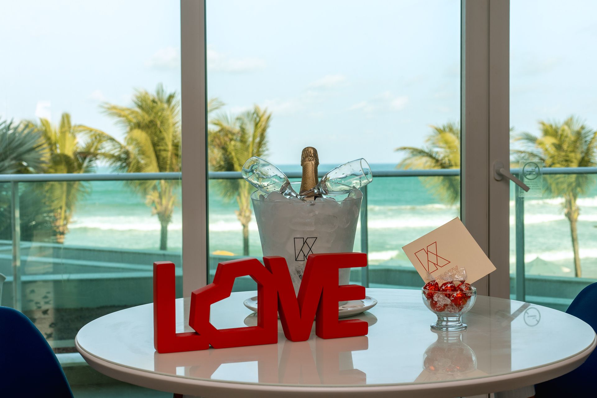 Mesa dentre de quarto de hotel, com balde de gelo e espumante dentro, taça de vidro com bombons de chocolate e um letreiro enfeitando a mesa escrito 