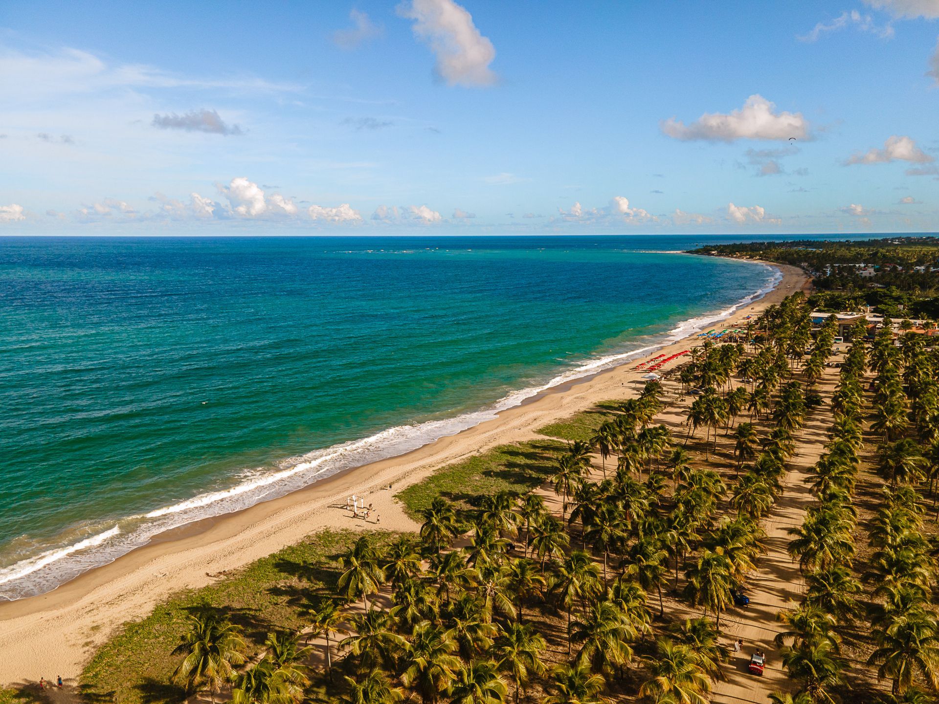 Uma visão aérea da praia, com uma grande faixa de areia e coqueiros. O mar tem uma coloração azul-turquesa e forma pequenas ondas que chegam à praia. O céu azul claro tem poucas nuvens esparsas.