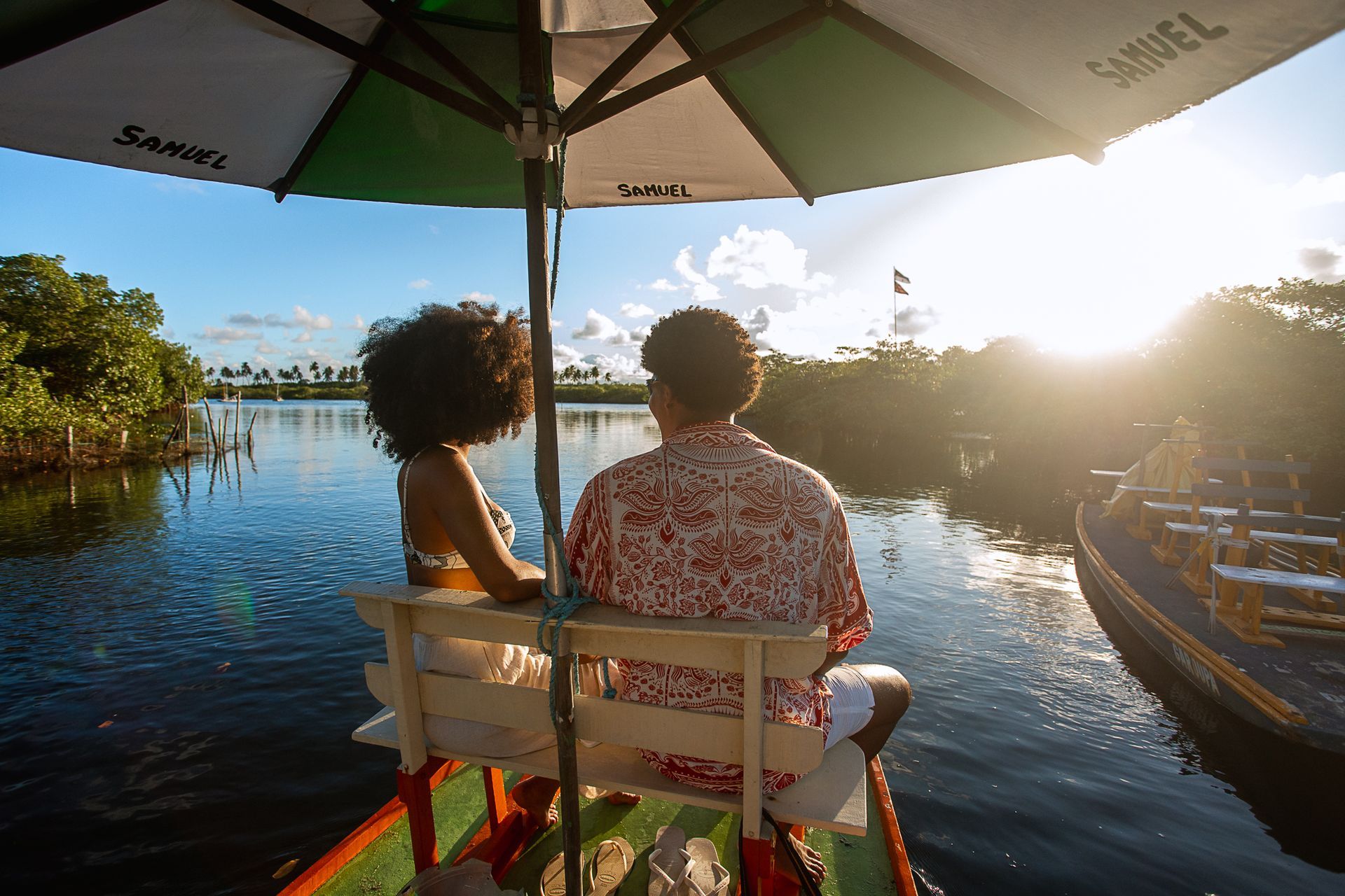 Casal sentado em banco na jangada, admirando o pôr do sol no mangue. A água é escura e estão cercados de mata nativa.