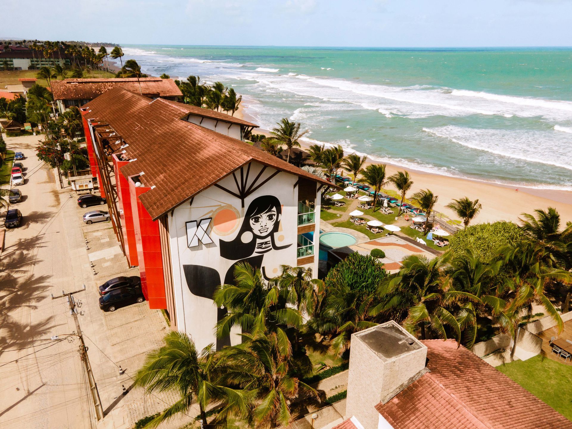 Vista aérea do Kembali: fachada com mural autoral, telhados coloniais, piscina e jardim de frente para o mar.