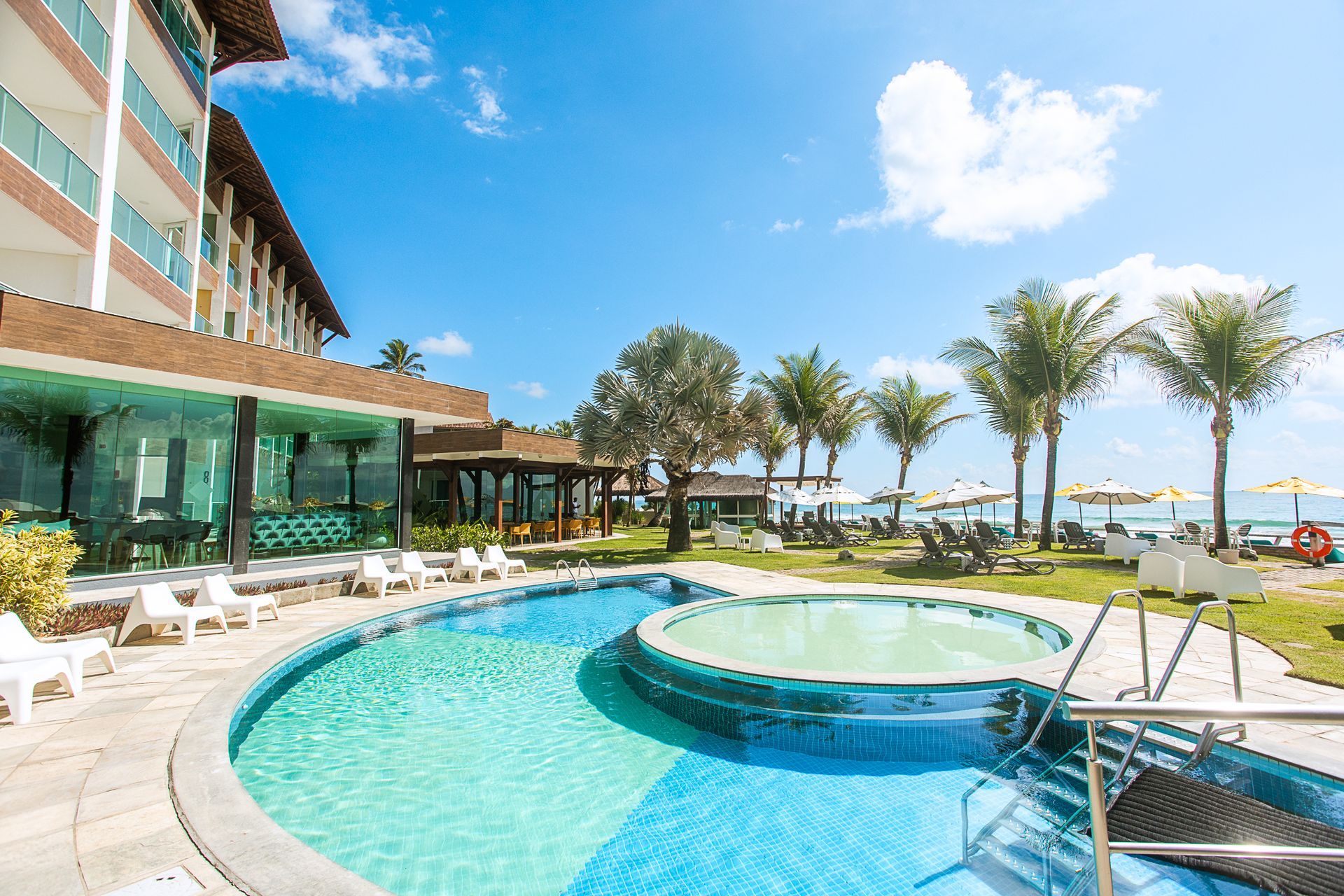Área externa do Kembali Hotel à beira-mar com piscina, espreguiçadeiras brancas, coqueiros e guarda-sóis amarelos em frente ao mar azul em um dia ensolarado.