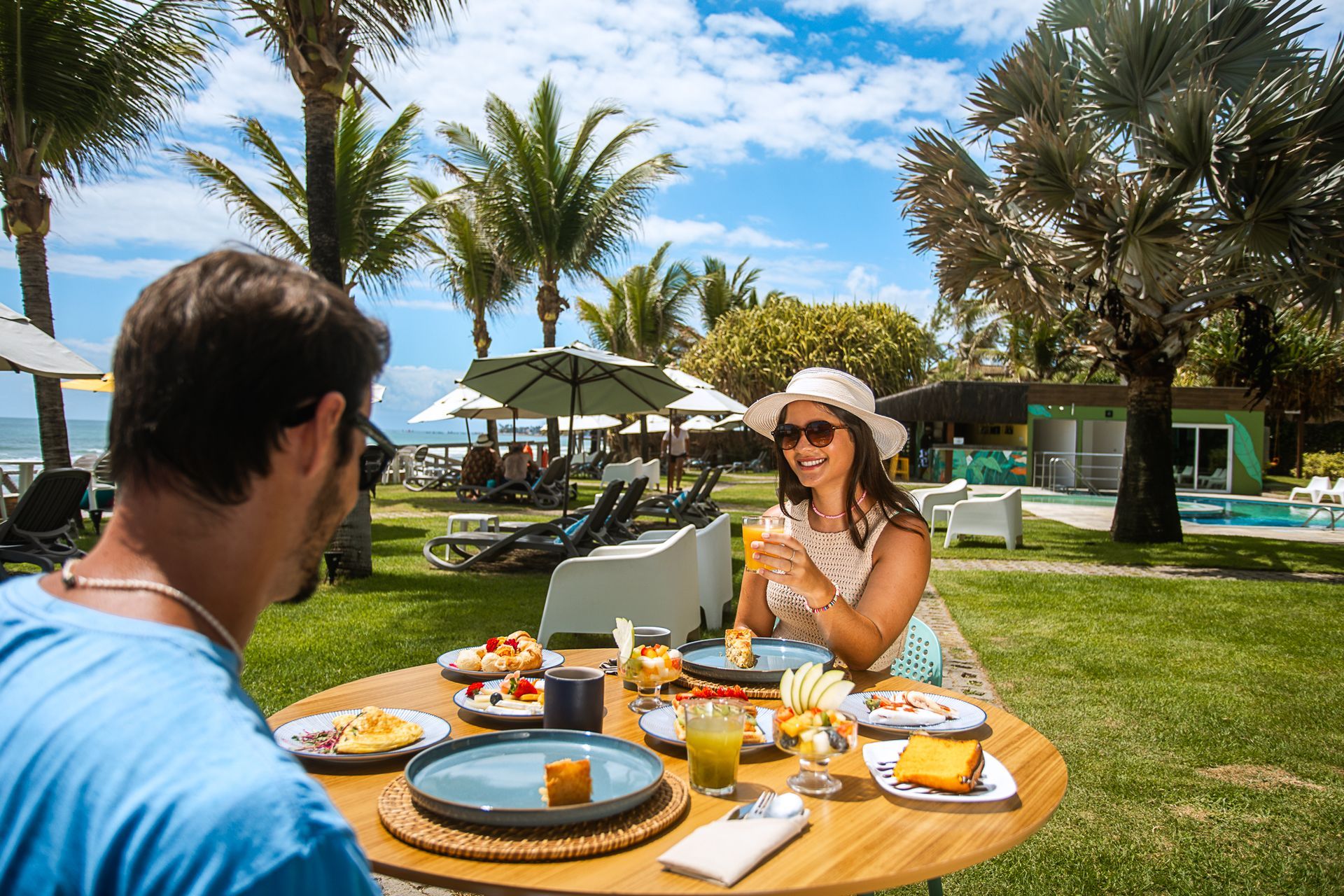 Casal aproveitando café da manhã ao ar livre em mesa posta com frutas, pães, sucos e outras opções, em área gramada com vista para o mar, piscina e coqueiros ao fundo em um dia ensolarado.