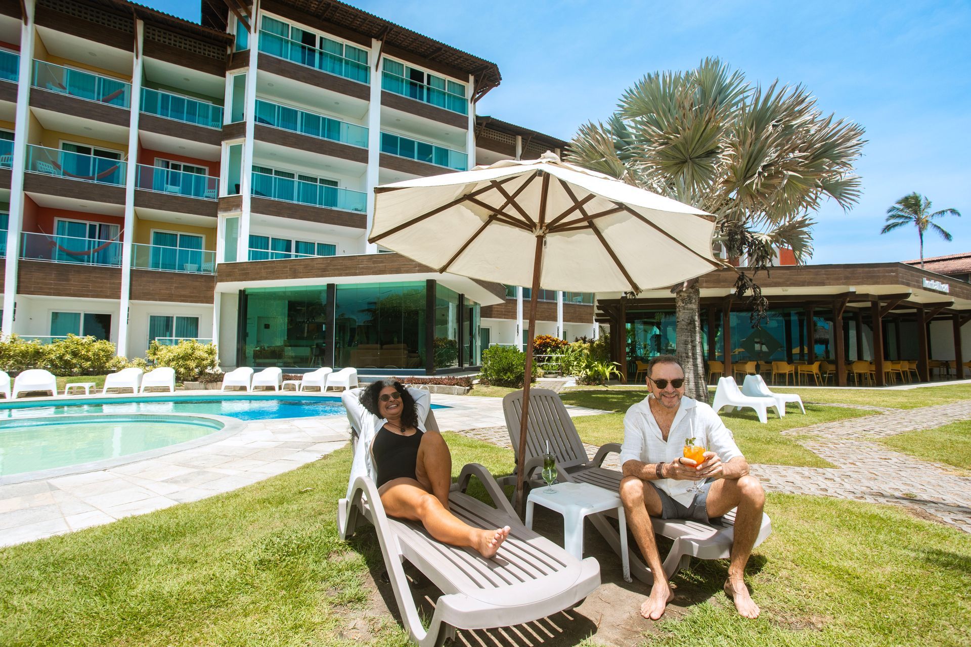 Casal relaxando em espreguiçadeiras à beira da piscina de hotel em Porto de Galinhas sob guarda-sol.