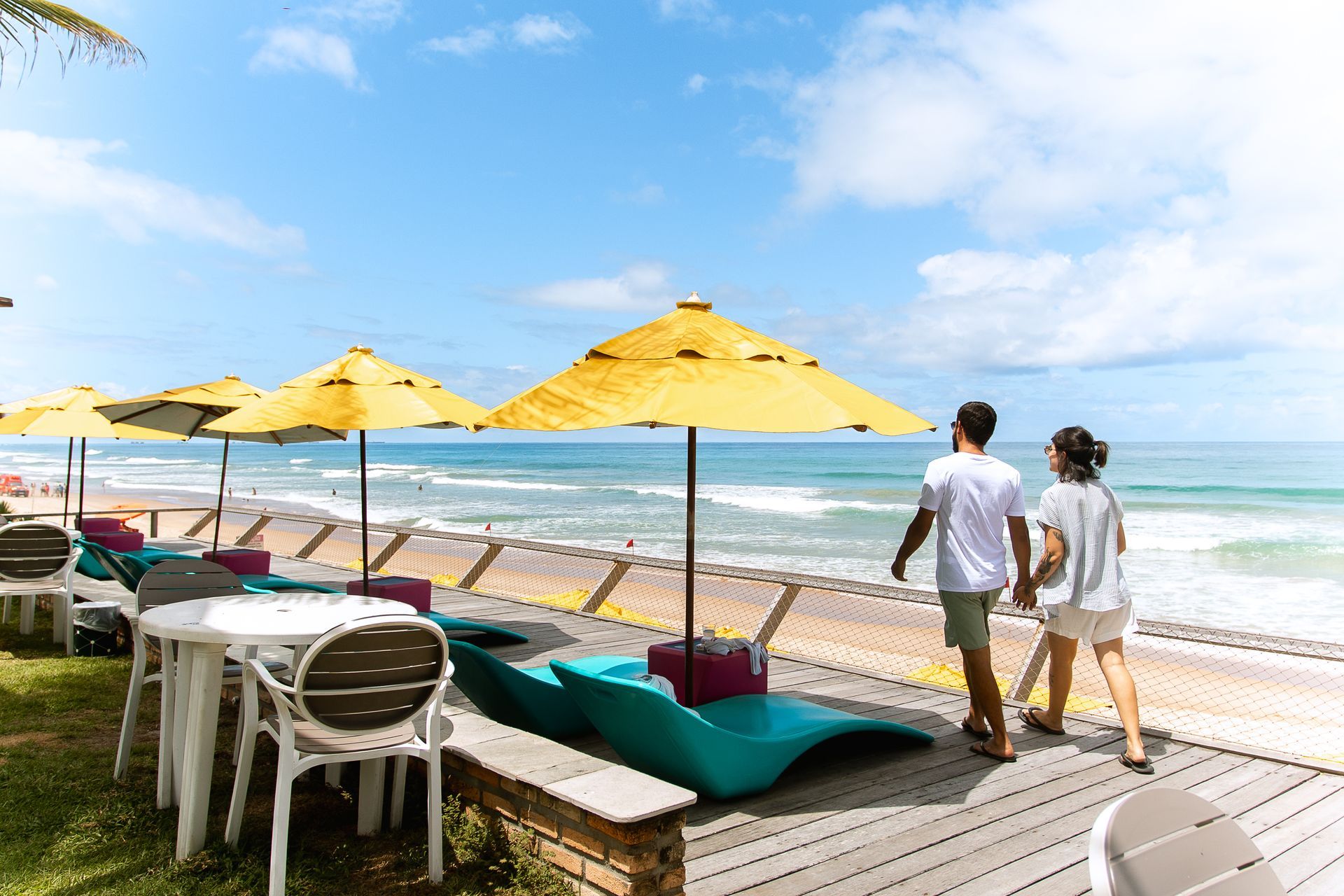 Casal caminha de mãos dadas no deck do Kembali Hotel. Espreguiçadeiras com guarda-sóis estão lado a lado, de frente para a praia. O mar está azul e o dia ensolarado.