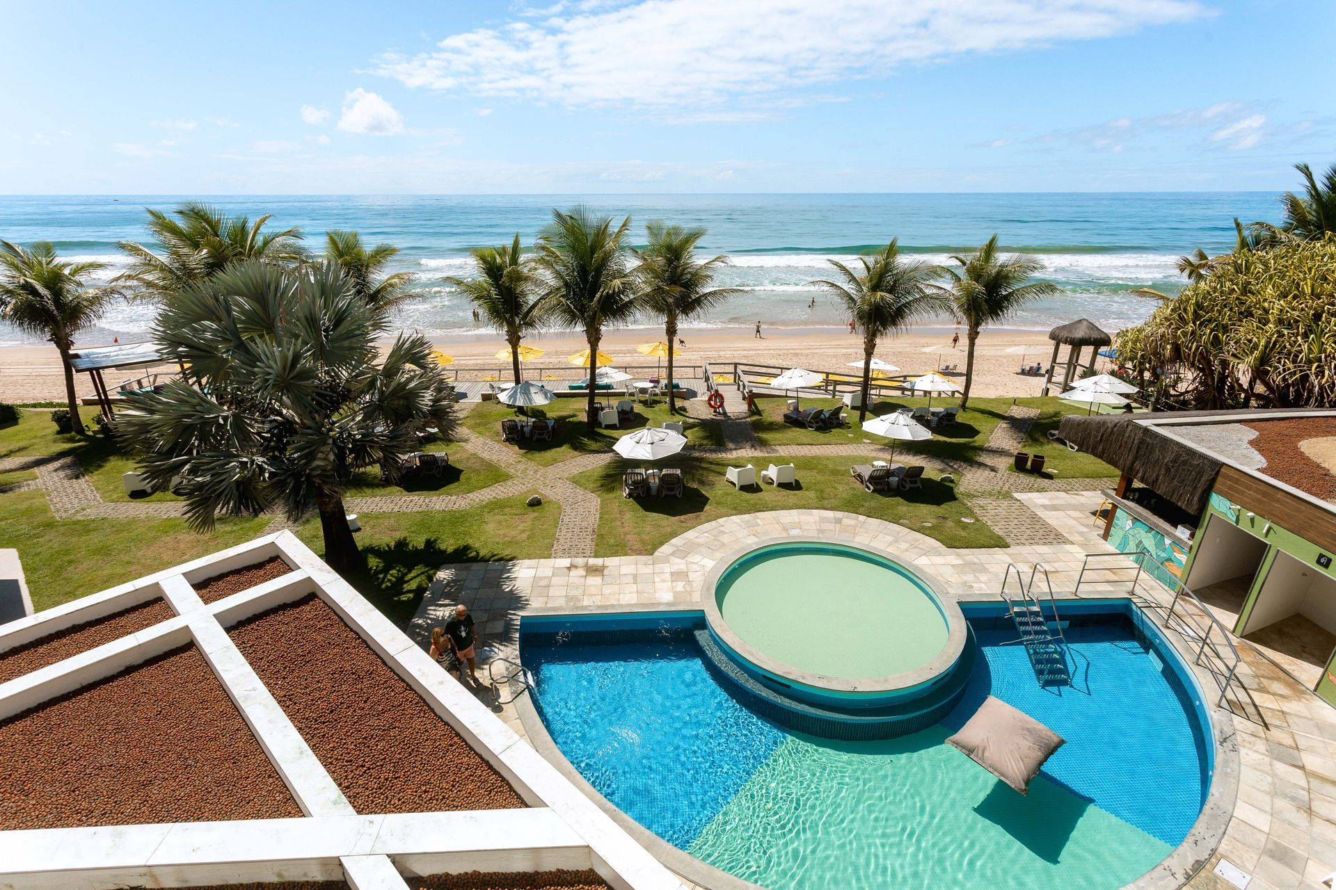 Vista aérea de hotel à beira-mar com piscina azul em primeiro plano, espreguiçadeiras e guarda-sóis distribuídos em um jardim com palmeiras. Ao fundo, a faixa de areia e o oceano com ondas suaves sob um céu claro e ensolarado.