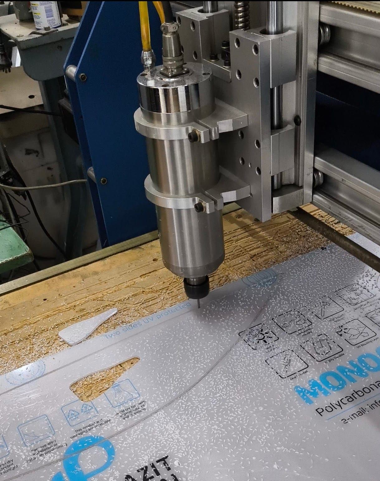 Máquina CNC cortando una lámina de plástico transparente; la máquina es plateada, la lámina está sobre una mesa de madera.