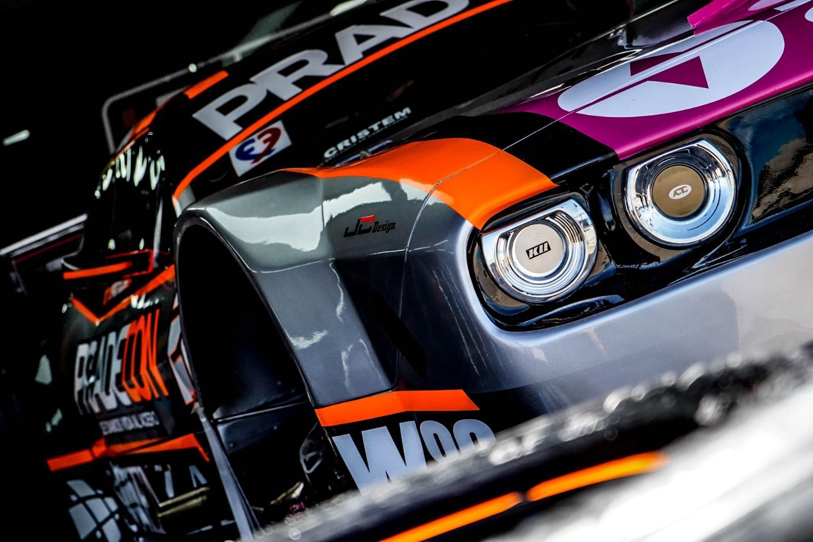 Coche de carreras, negro, naranja y morado, con logotipos de patrocinadores y faros cromados brillantes.