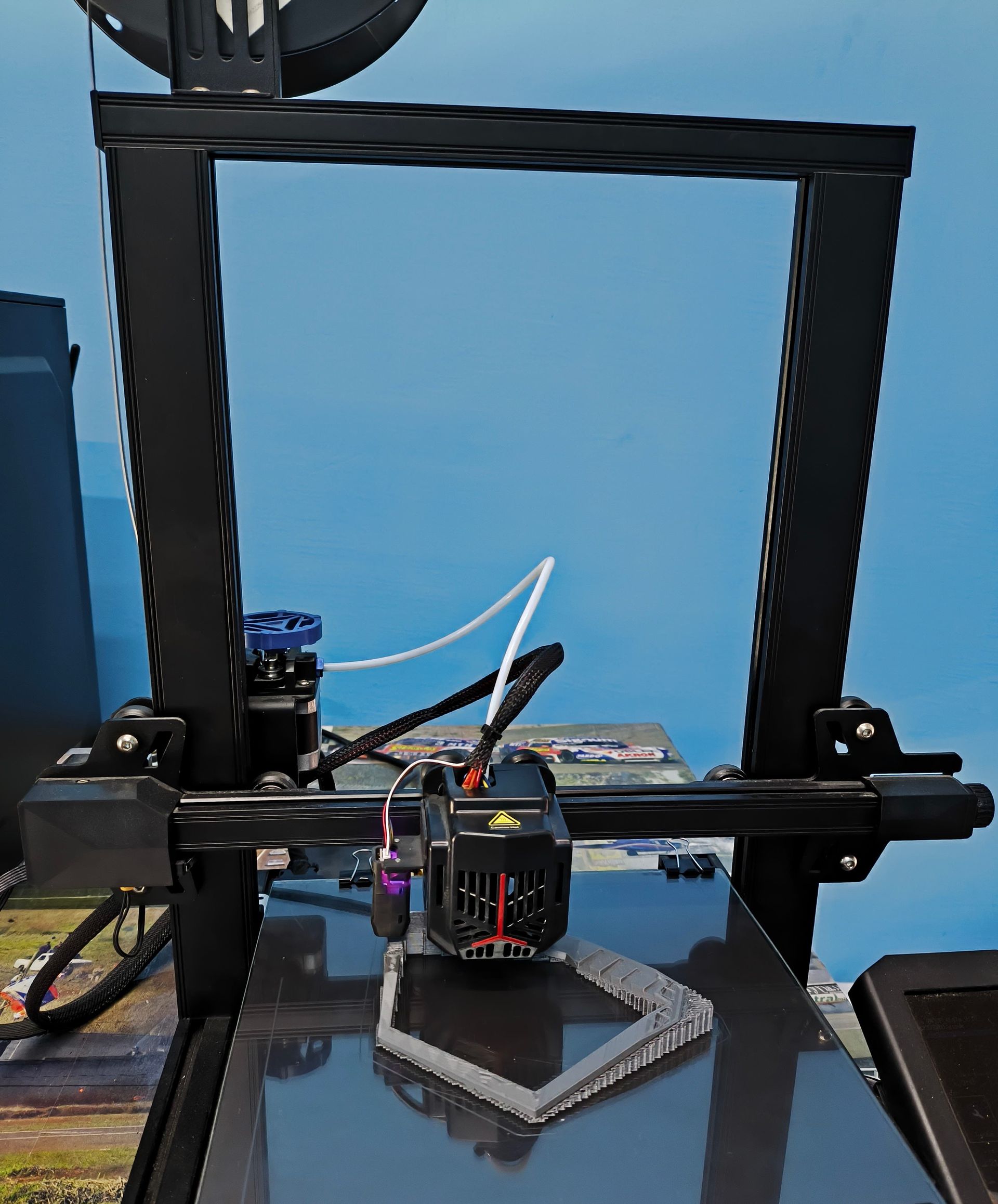 Impresora 3D en funcionamiento, imprimiendo una forma gris sobre una base negra. Fondo de cielo azul.