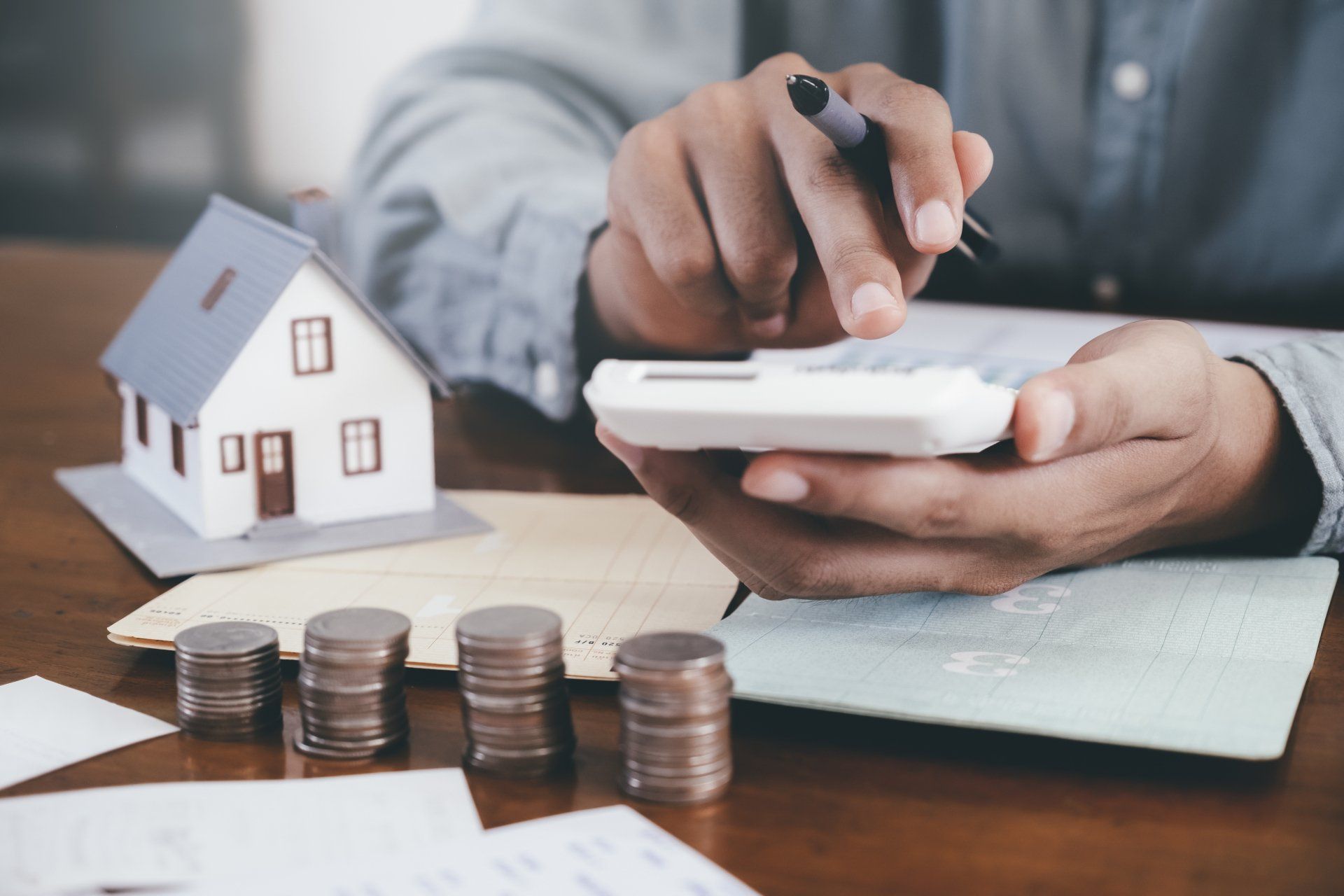 Person calculating finances with a model house, coins, and documents on a table.