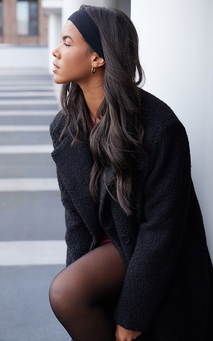 Mujer vestida con un traje oscuro, apoyada contra un pilar blanco en un estacionamiento.