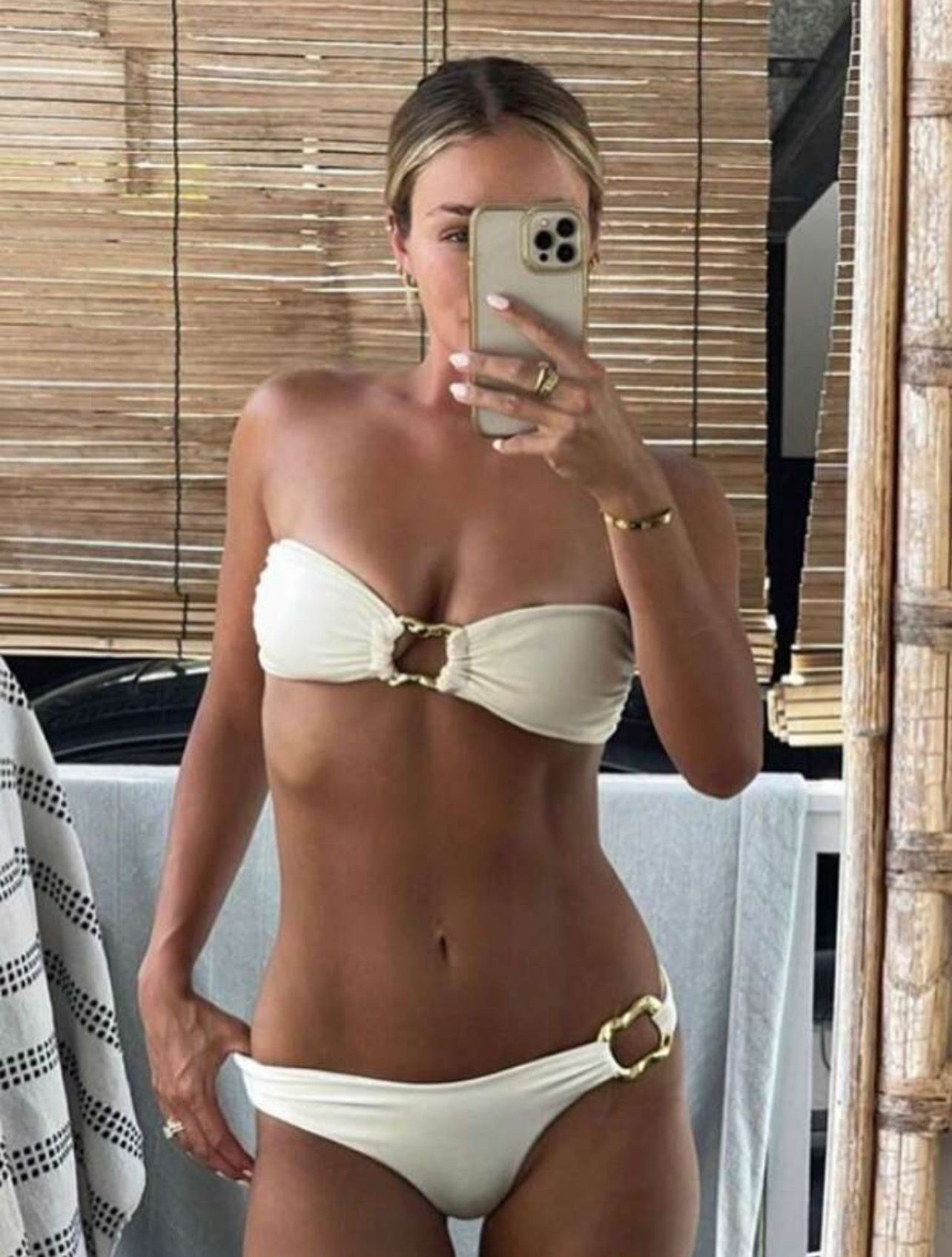 Mujer en bikini blanco, top con cuello halter, de pie en el interior, cerca de una toalla verde y blanca.