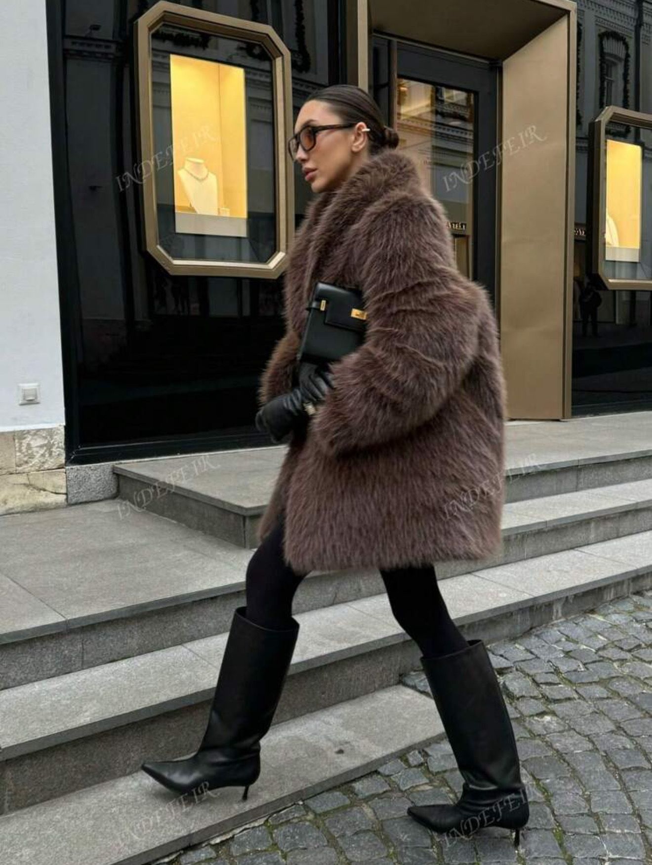 Mujer con un abrigo de piel marrón, gafas de sol y guantes negros, de pie frente a una tienda.