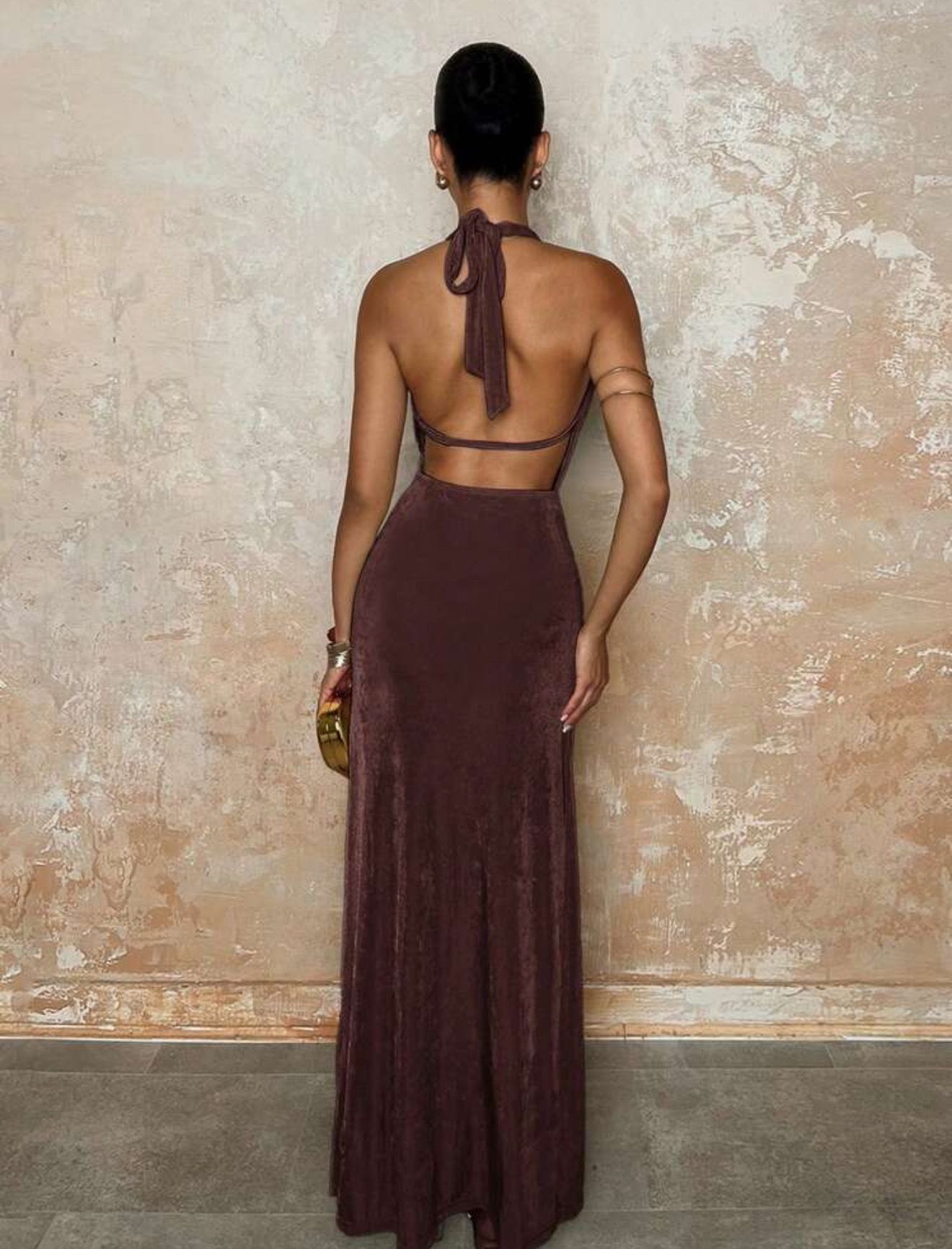 Mujer con un vestido halter marrón oscuro con una abertura alta, sosteniendo un clutch dorado y usando tacones dorados.