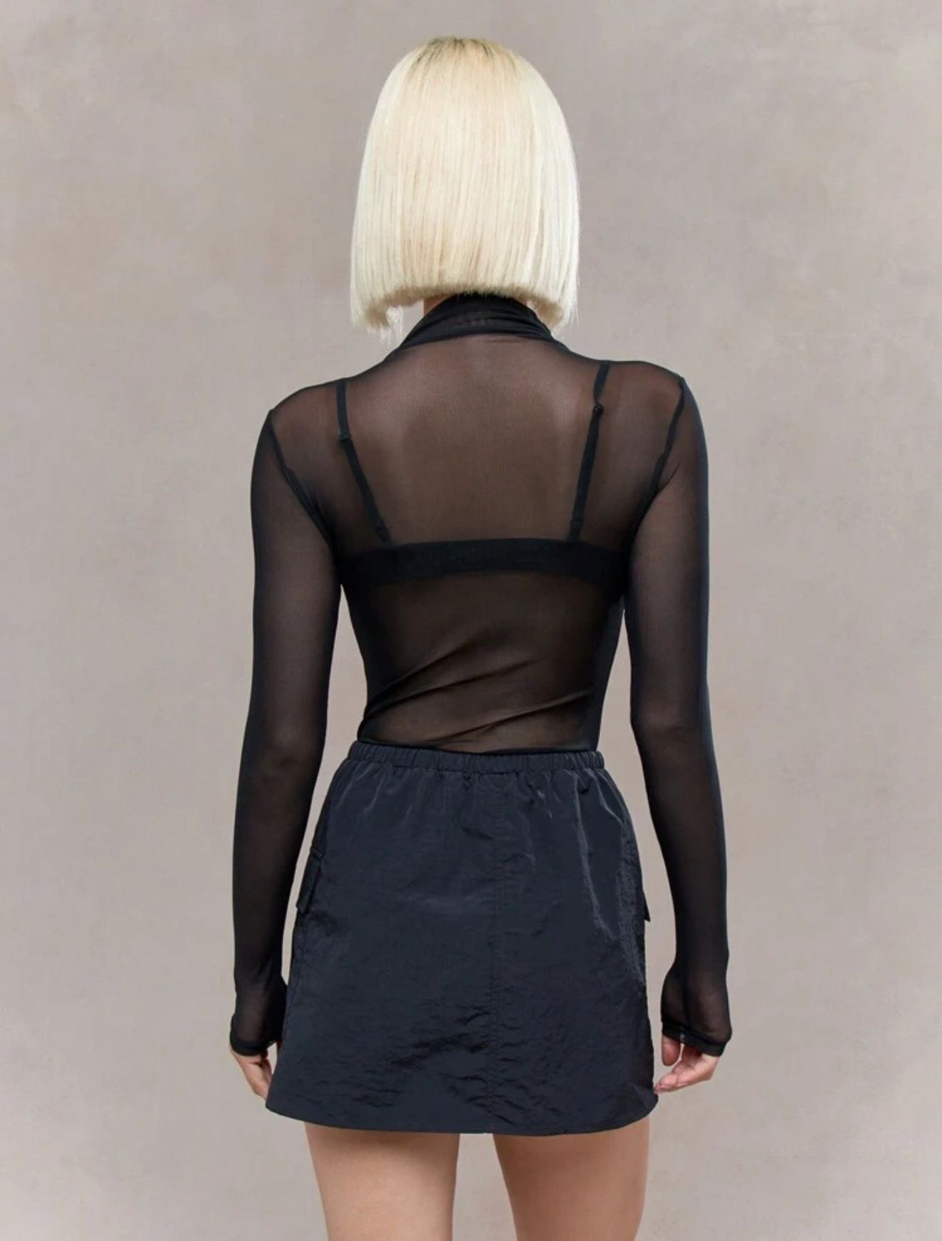 Mujer con top de malla negra, sujetador y falda, mirando hacia la izquierda, con las manos cerca del cuello.