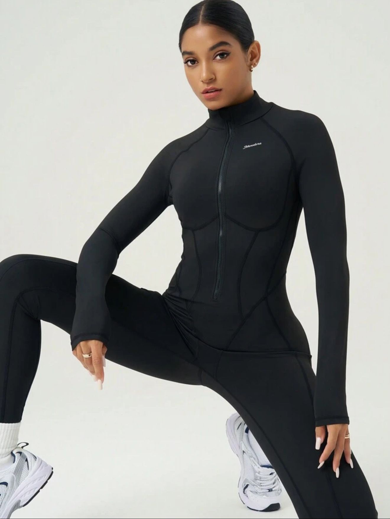 Mujer con mono negro, sosteniendo un bolso y una botella de agua, con gafas de sol, de pie en el estudio.