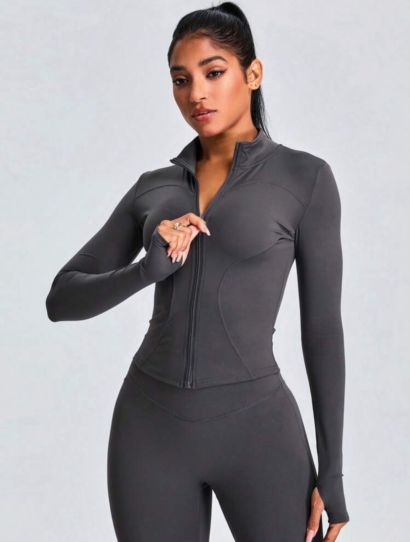 Mujer con conjunto deportivo gris, chaqueta con cremallera, leggings y zapatillas deportivas, posando sobre un fondo gris.