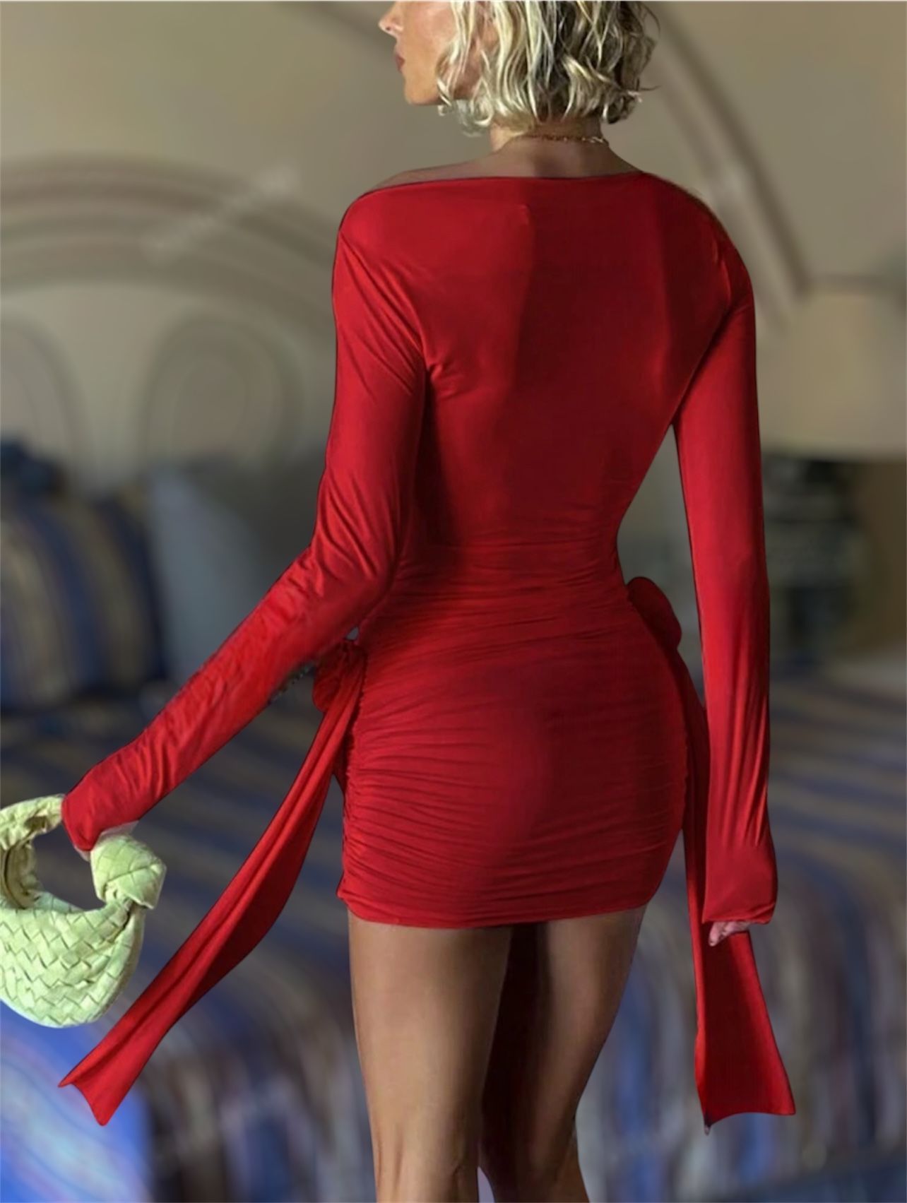 Mujer con un minivestido rojo fruncido, sosteniendo un bolso claro. Al aire libre, frente a un edificio blanco.