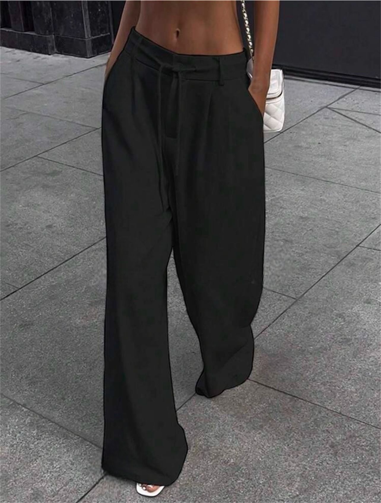 Persona con pantalones negros de pierna ancha y manos en los bolsillos; bolso blanco; entorno al aire libre.