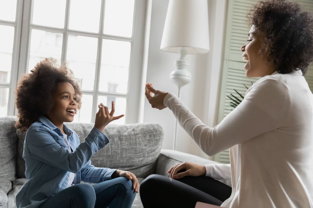 Woman and child smiling, using sign language in a bright living room with a window and lamp.