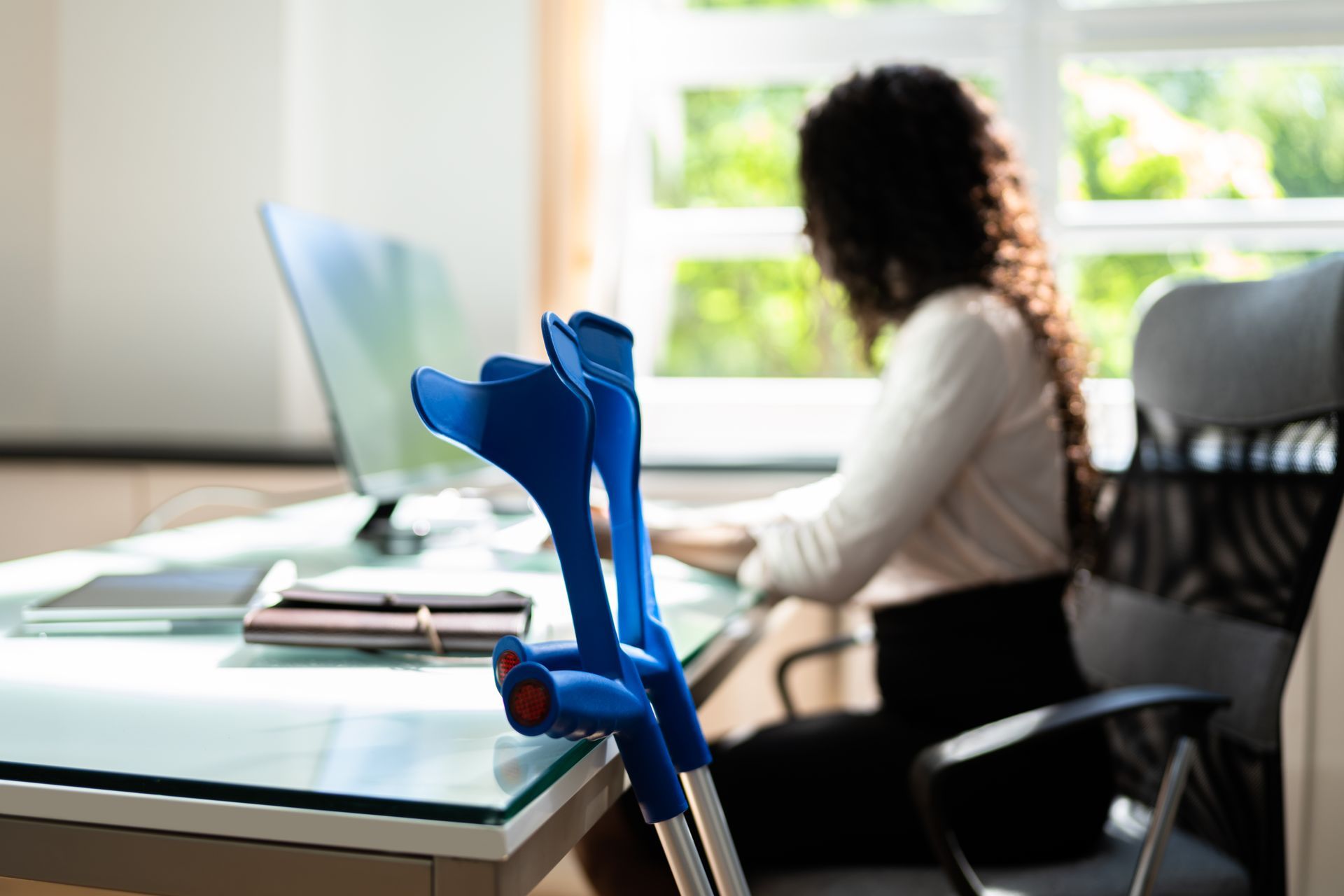 Blue crutches in focus on a desk; a person works on a computer in the background.