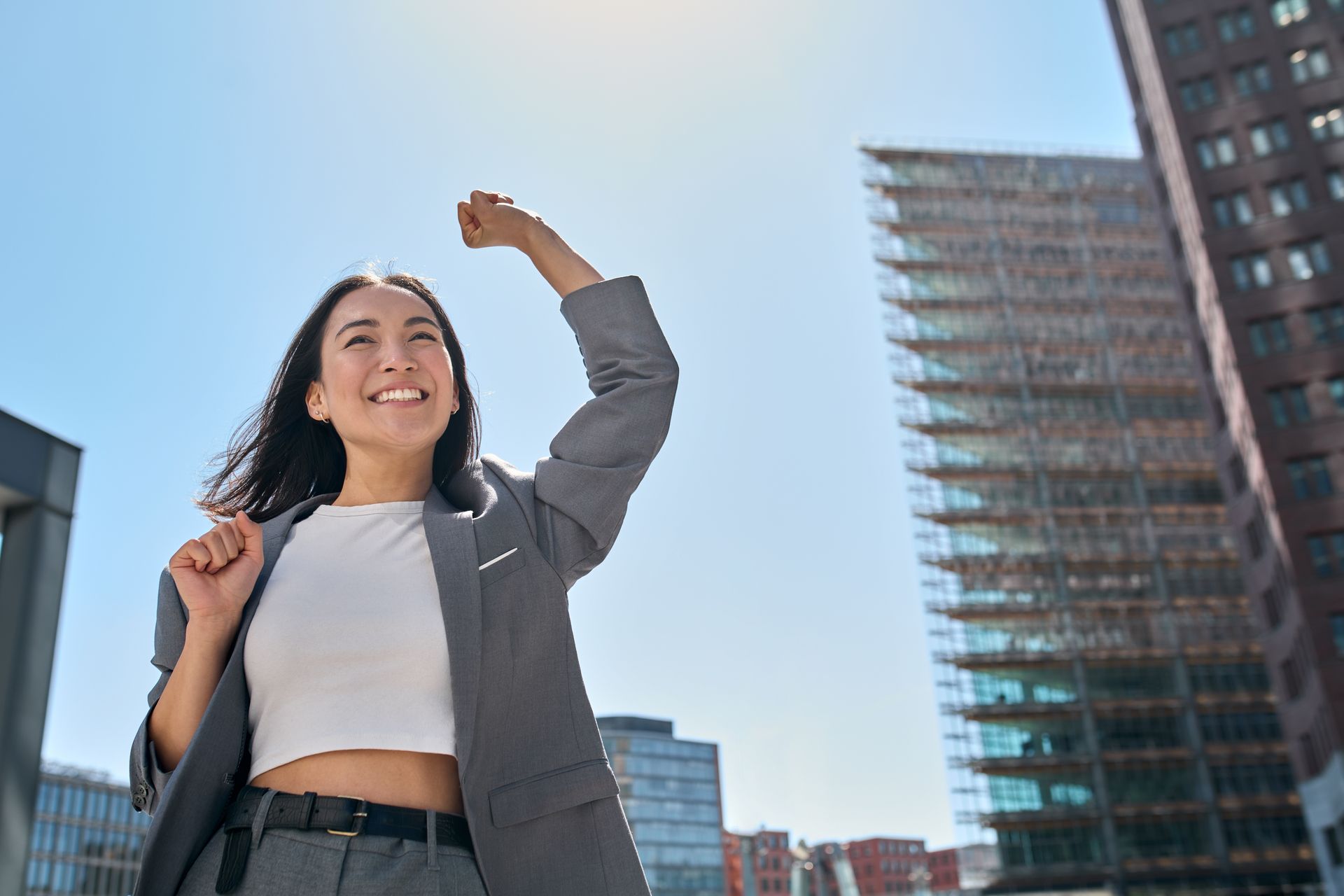 Woman with fist raised in front of city buildings, smiling, wearing blazer and white top.