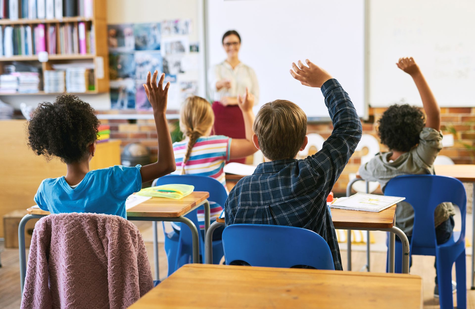 Students raising hands in a classroom, teacher at whiteboard in background.