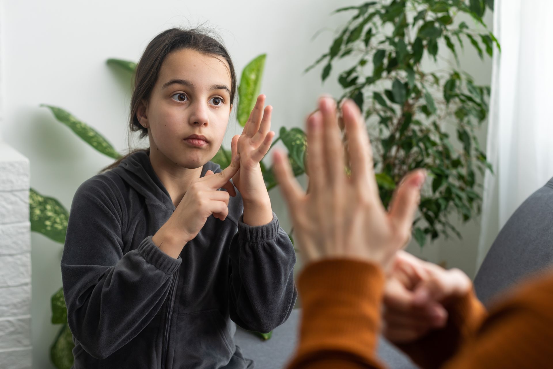 Two people using sign language indoors, with green plants in the background.