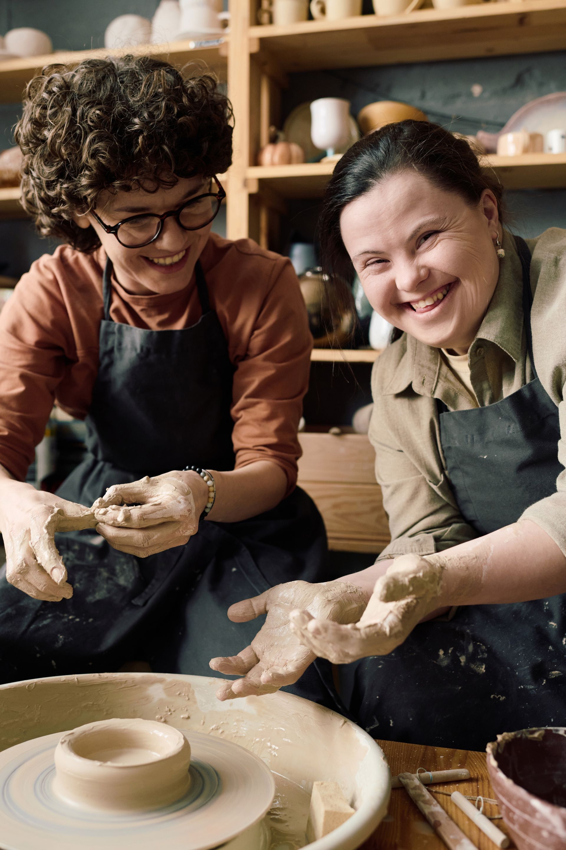 Two people smile while working with clay at pottery wheels. They wear aprons in a workshop setting.