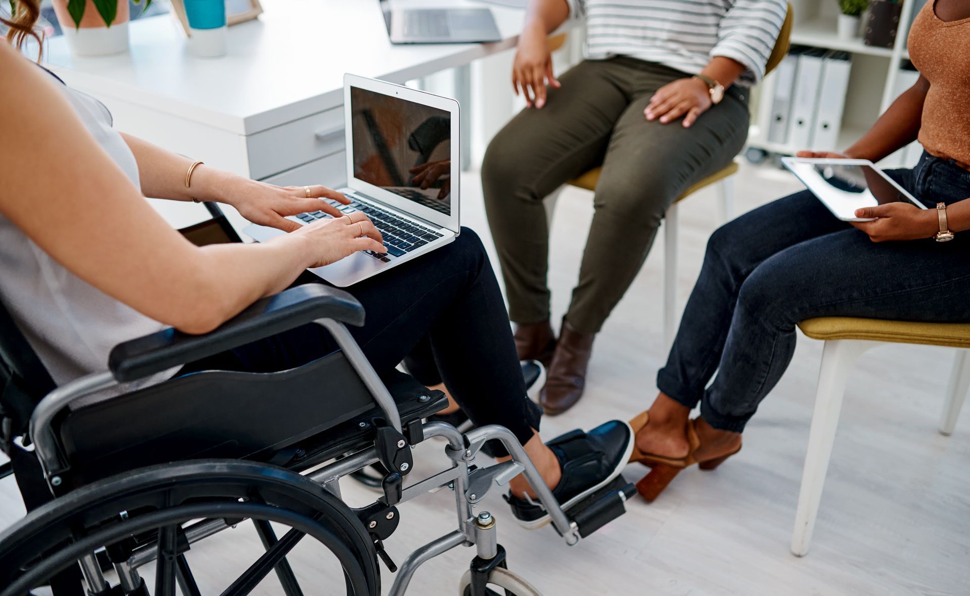 Three people in a meeting. One uses a wheelchair and laptop, another has a tablet, and the third watches.