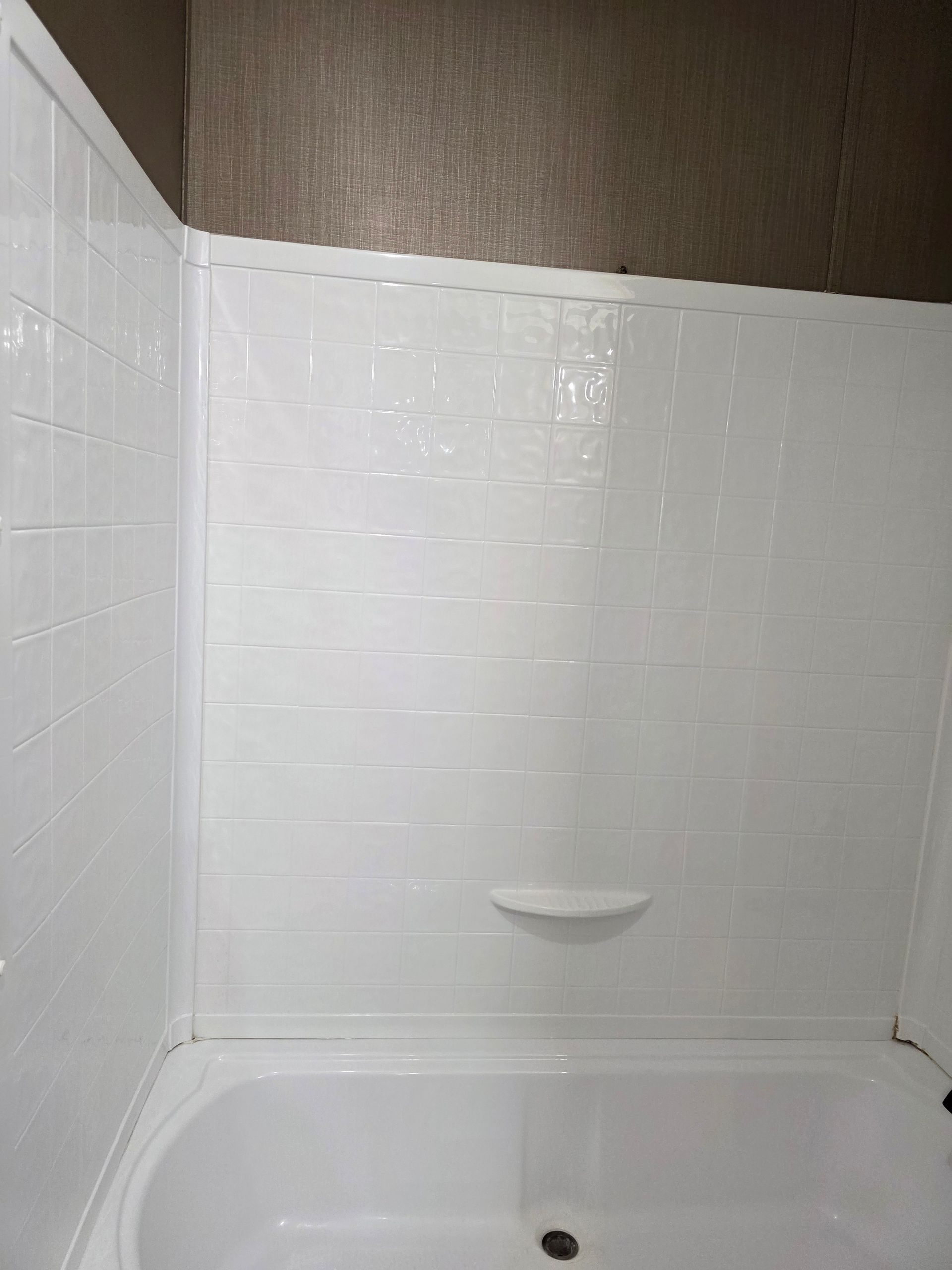 White shower stall with built-in soap dish, bathtub, and textured brown wall above.