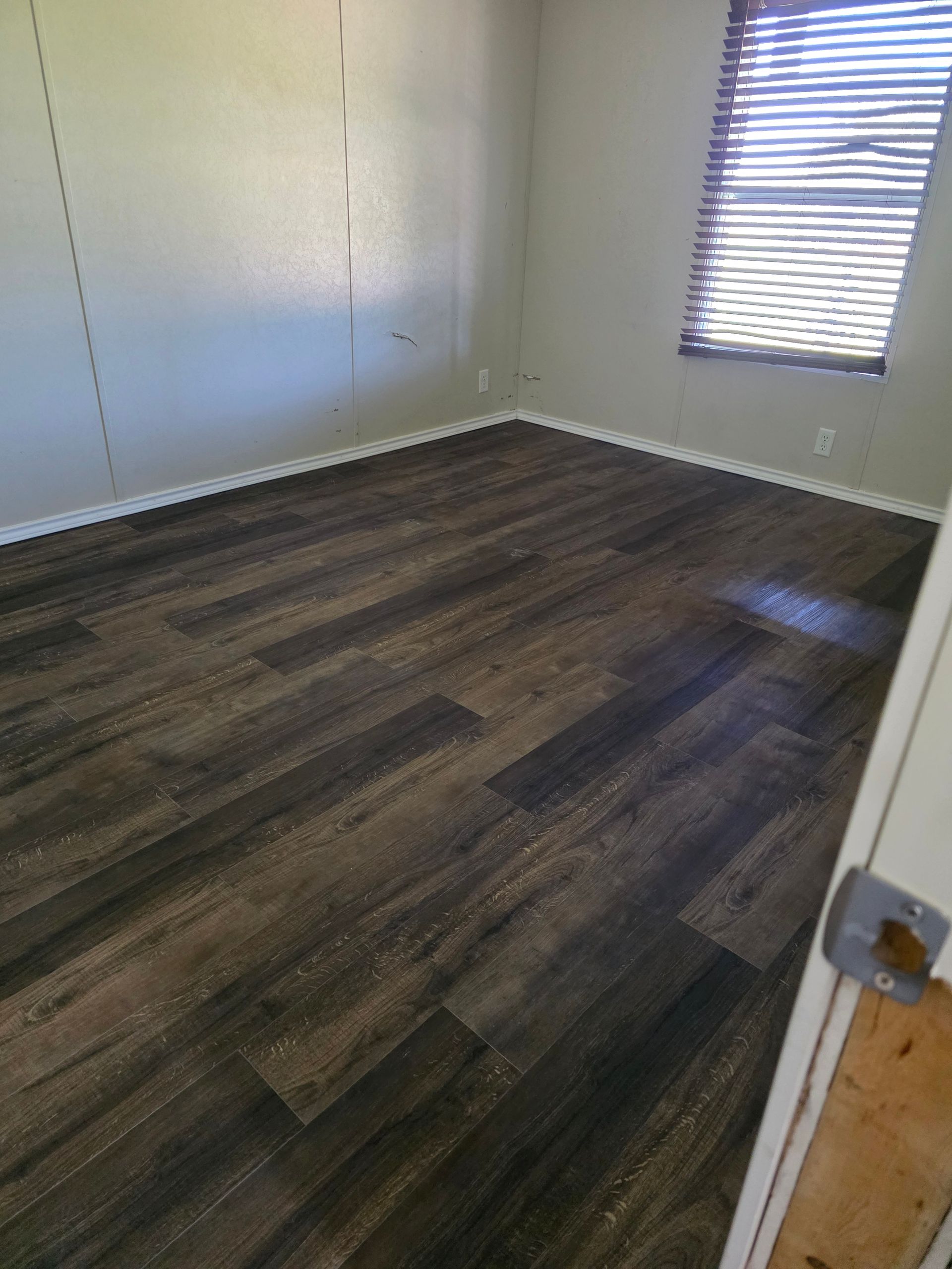 Empty room with new, dark wood-look flooring and a window with blinds. Beige walls, white trim.